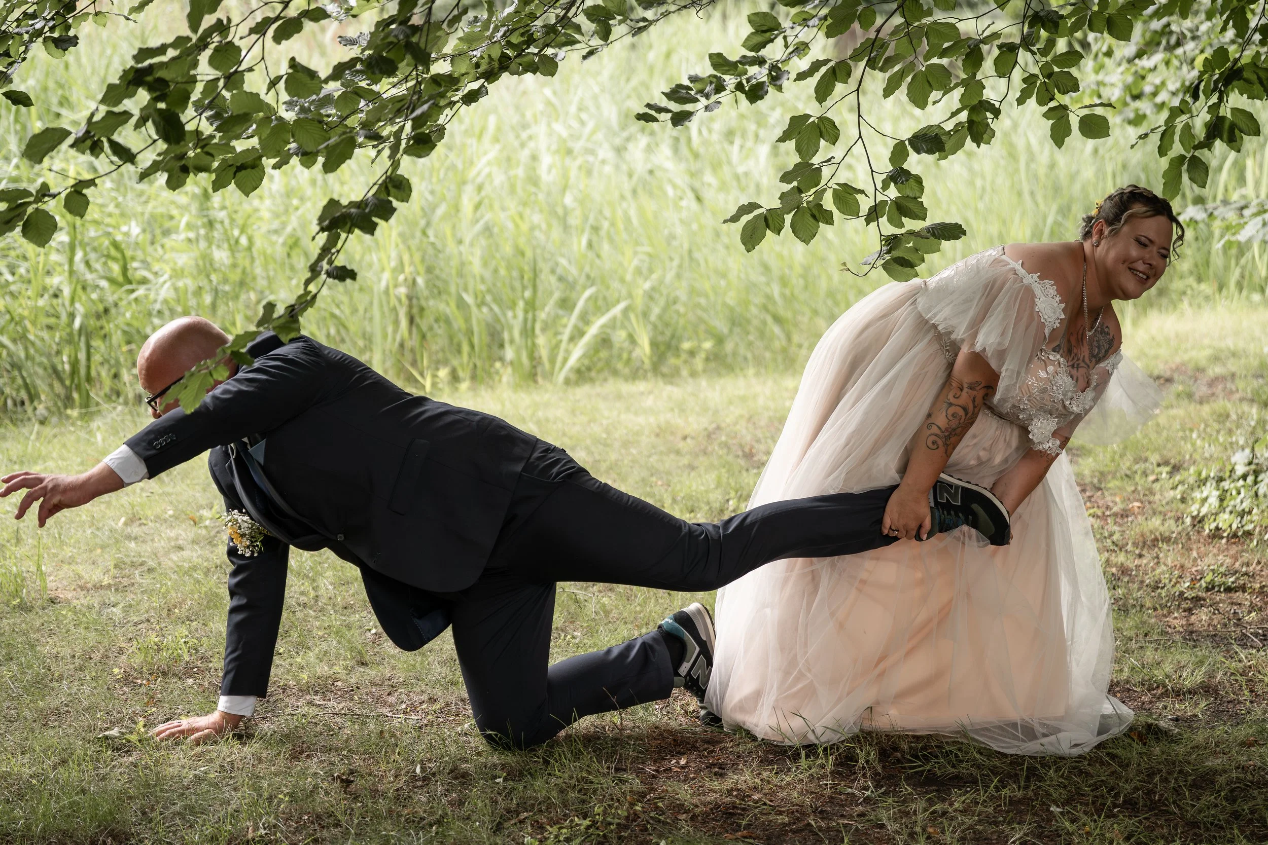 Ein Mann in Anzug und eine Frau im Hochzeitskleid stehen in einem Wald, die Frau ist lachend, während der Mann in einem knienden Würgegriff gehalten wird, alle auf grasbewachsenem Boden unter grünem Baum.