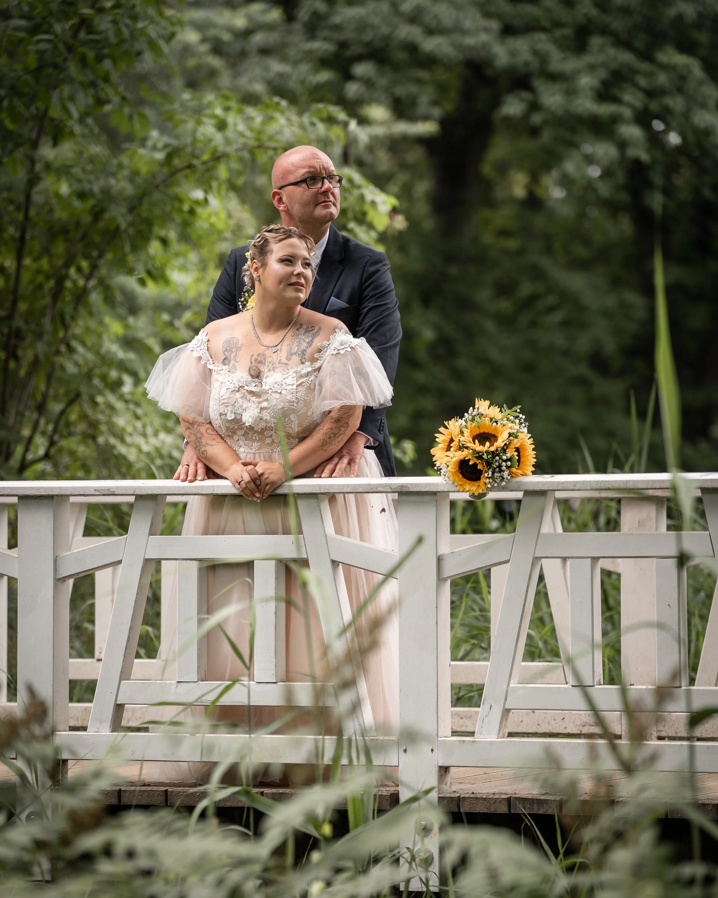 Ein Mann und eine Frau stehen auf einer Brücke in einem grünen Garten, die Frau trägt ein weißes Kleid mit Spitze und Tattoos auf Armen und Dekolleté, die Frau hält eine Sonnenblume.