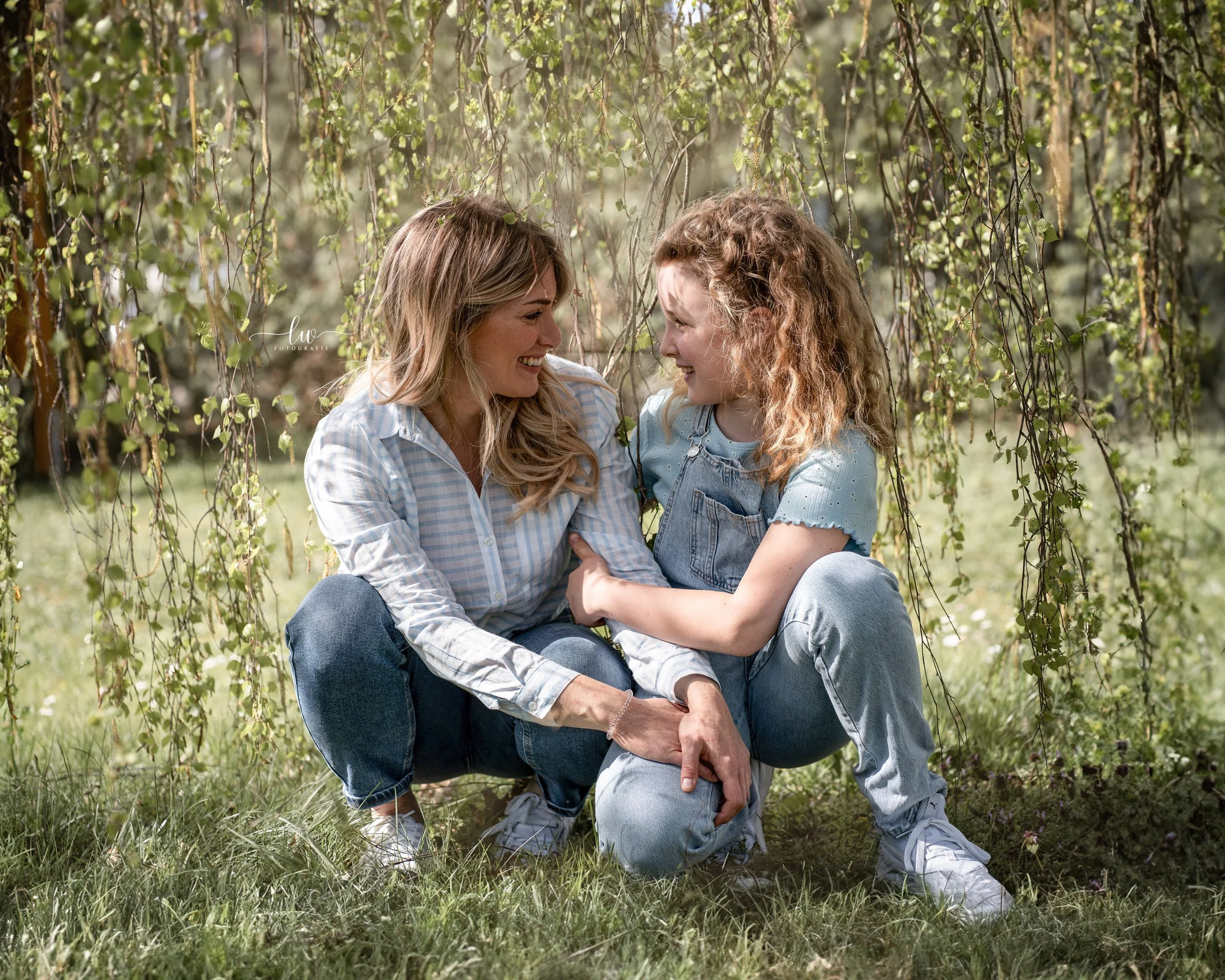 Zwei Frauen sitzen im Grünen unter einem Baum, lachen sich an und schauen sich freudig an, während sie sich umarmen und sich gegenüber sitzen.