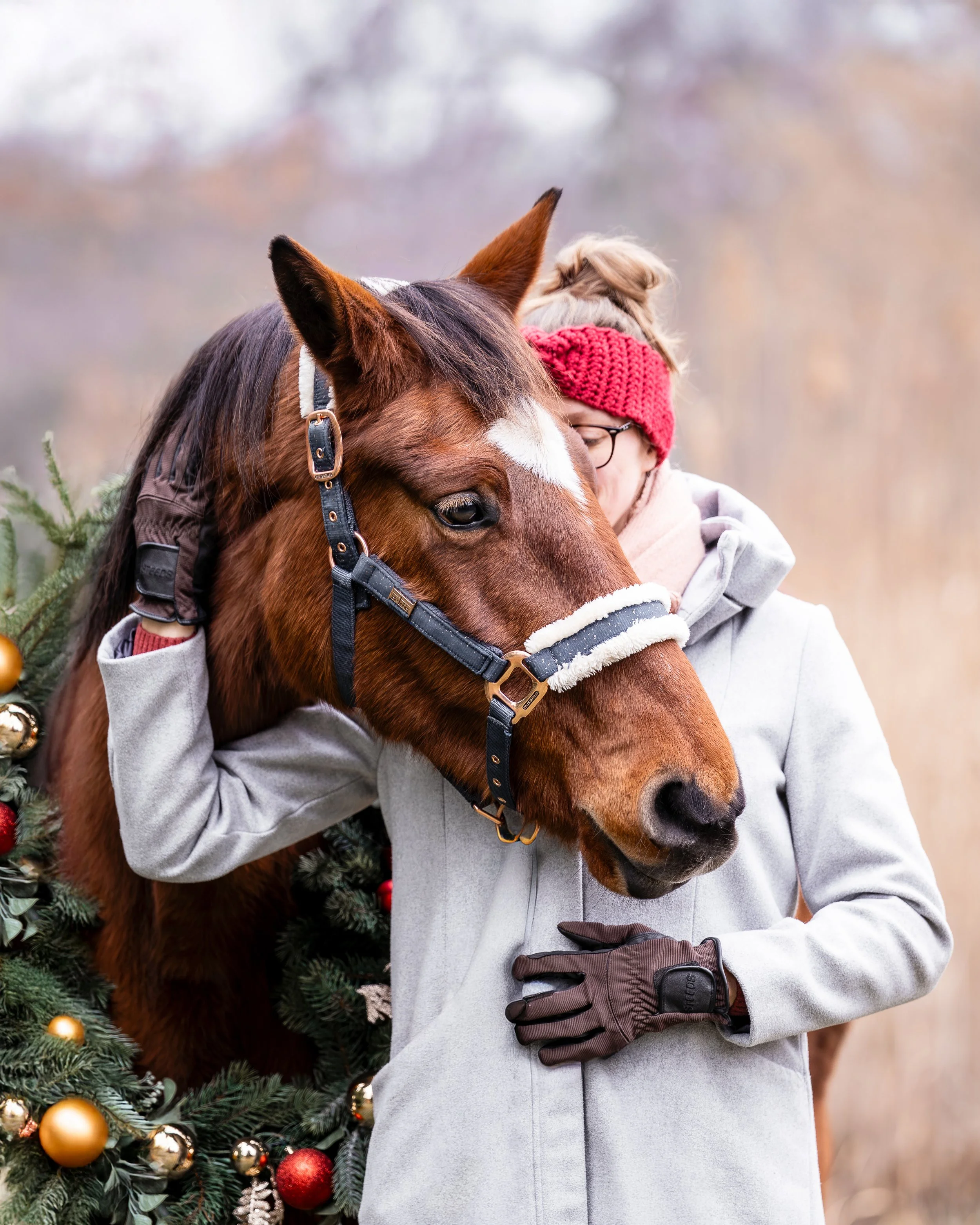 Ein Kind umarmt ein braunes Pferd mit weißer Markierung im Gesicht, im Hintergrund Weihnachtsbaum mit goldenen und roten Kugeln, trägt eine rote Wollmütze und graue Jacke.