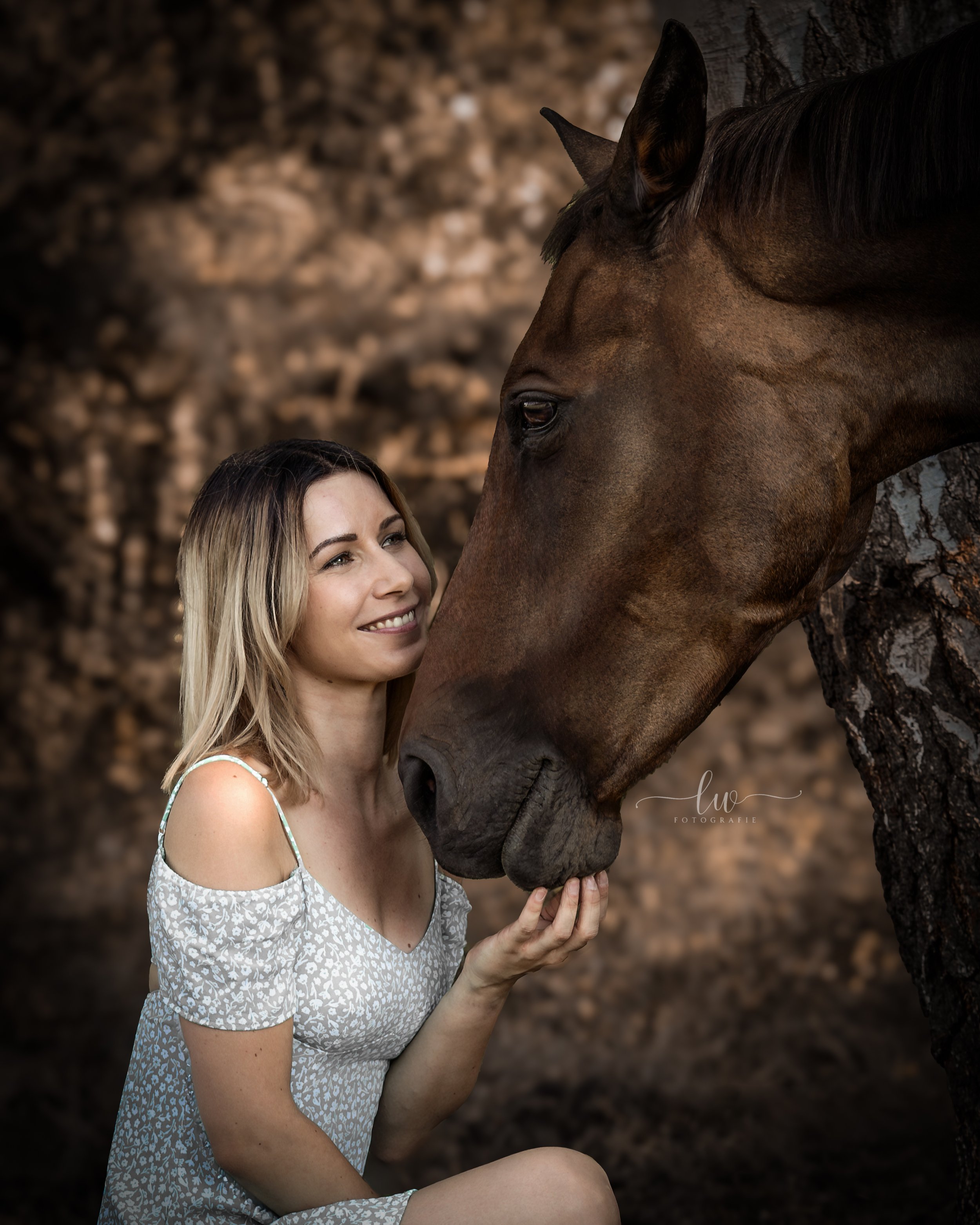 Eine lächelnde Frau mit blonden Haaren, die ein weißes Kleid mit einem floralem Muster trägt, streichelt ein braunes Pferd mit dunklem Mähne in einer natürlichen, bewaldeten Umgebung.