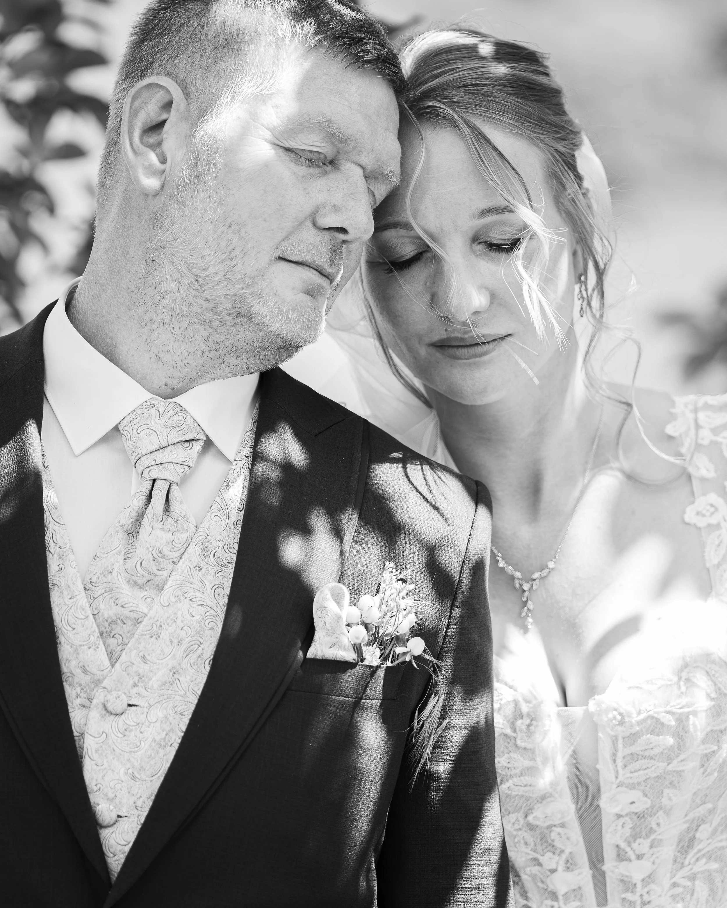 Das Bild zeigt ein Paar bei einer Hochzeitszeremonie, beide mit geschlossenen Augen und in einer innigen Umarmung. Die Frau trägt ein Brautkleid und eine Halskette, der Mann trägt einen Anzug mit Weste und Krawatte. Sie scheinen einen emotionalen Mom