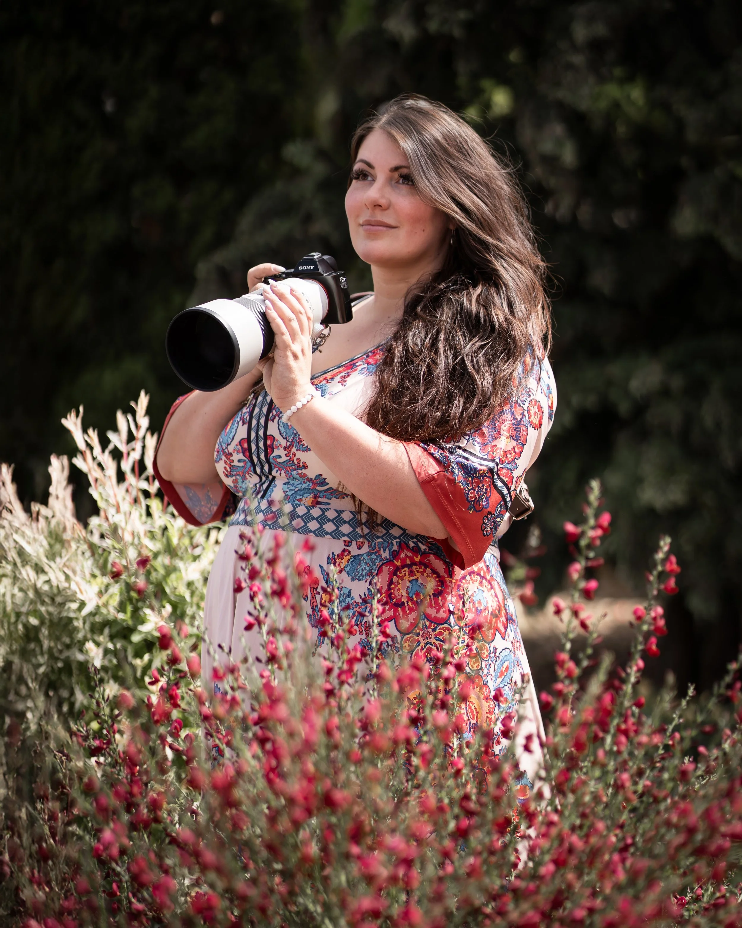 Eine Frau mit braunen, welligen Haaren, die ein Kameraobjektiv hält, steht in einem Garten mit blühenden Blumen und dunklem Hintergrund, lächelt leicht und schaut in die Ferne.