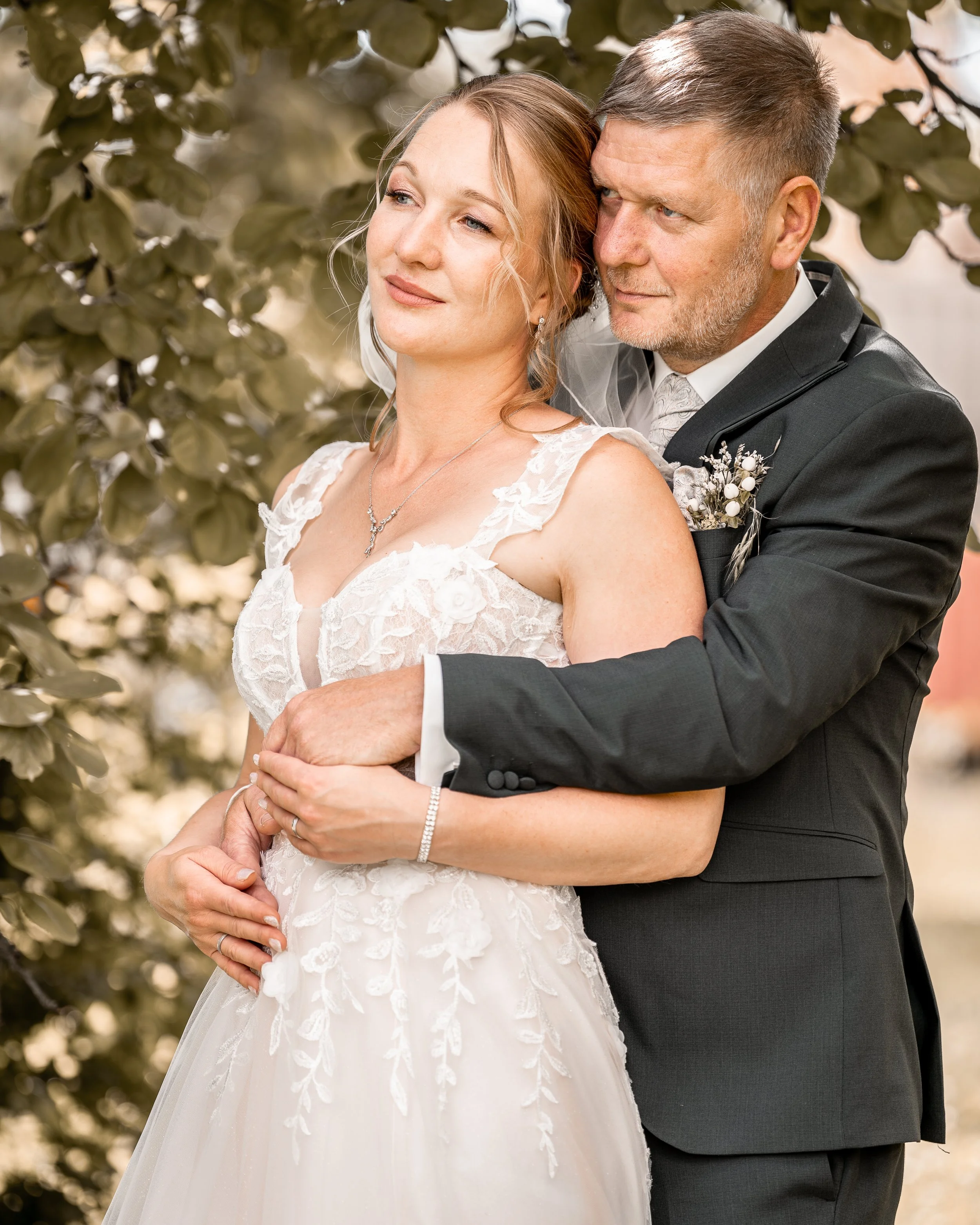 Ein Paar bei einer Hochzeit, die Frau trägt ein weißes Brautkleid mit Blumenmuster und der Mann trägt einen schwarzen Anzug. Sie stehen nah beieinander im Freien, umgeben von grünen Blättern.