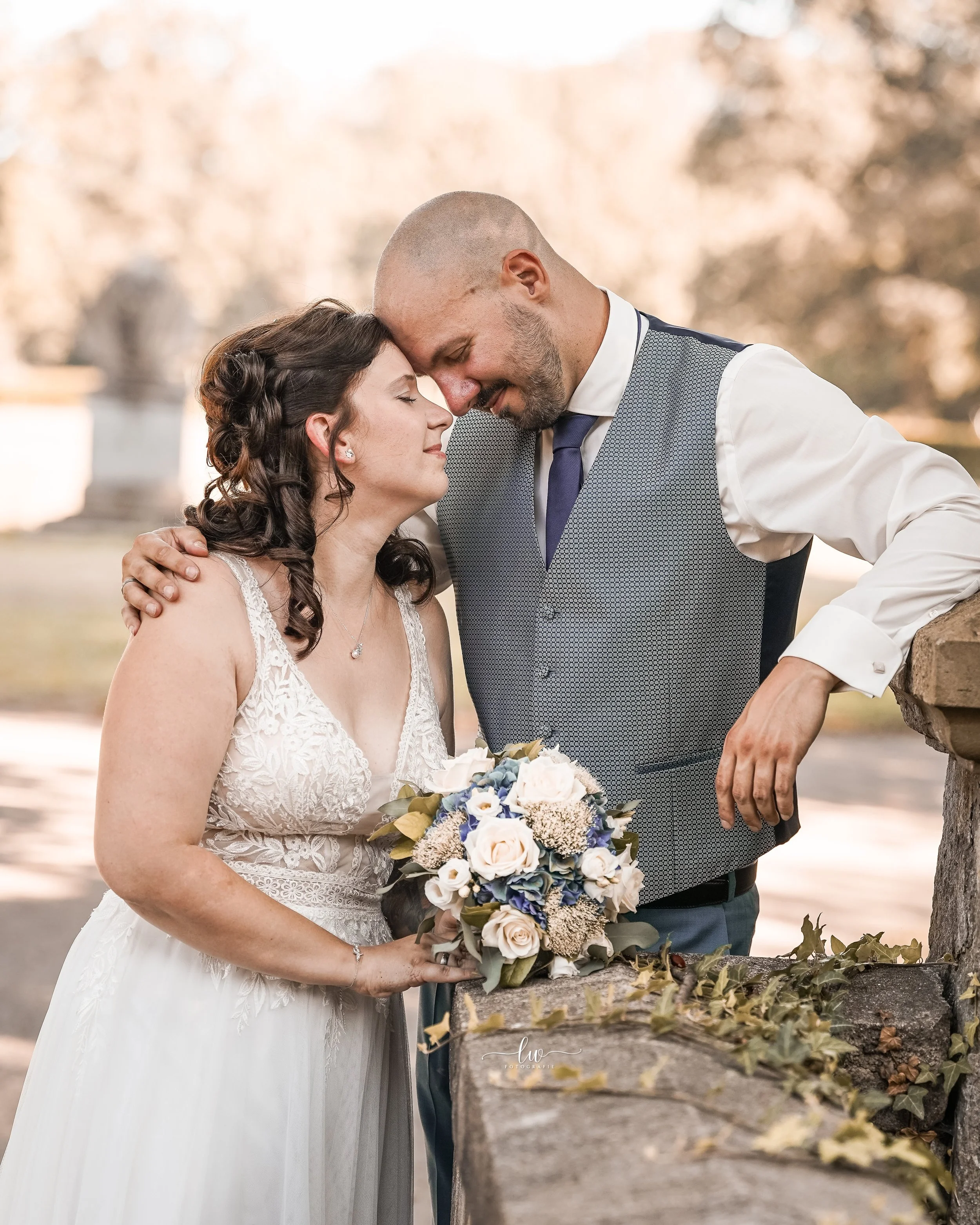 Ein heiratswilliges Paar, das sich an einem sonnigen Tag in einem Park umarmt und küsst, mit einer Braut in weißem Kleid und einem Bräutigam in Anzug mit Weste, im Hintergrund Bäume und eine Mauer.