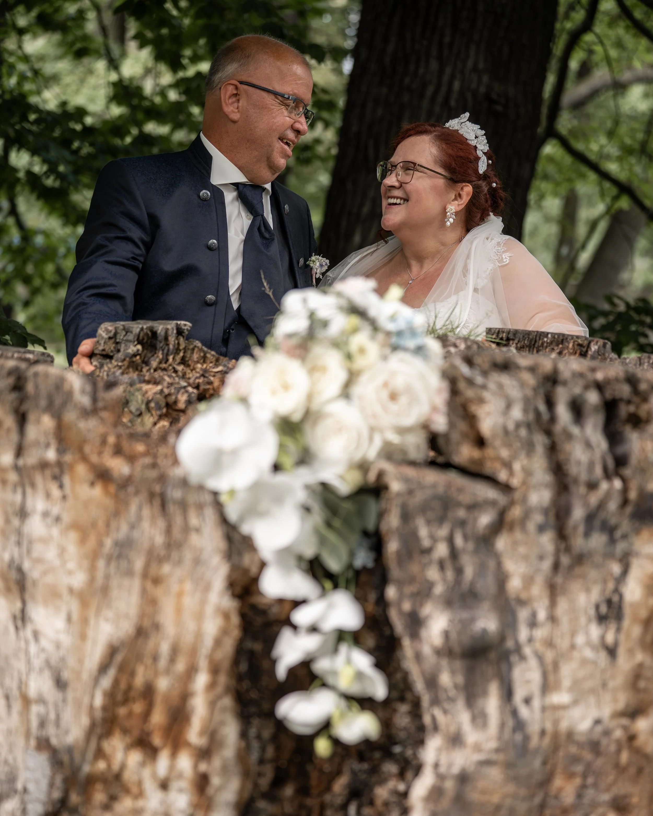 Ein älteres Paar in formeller Kleidung bei einer Hochzeit im Freien, umgeben von Bäumen.