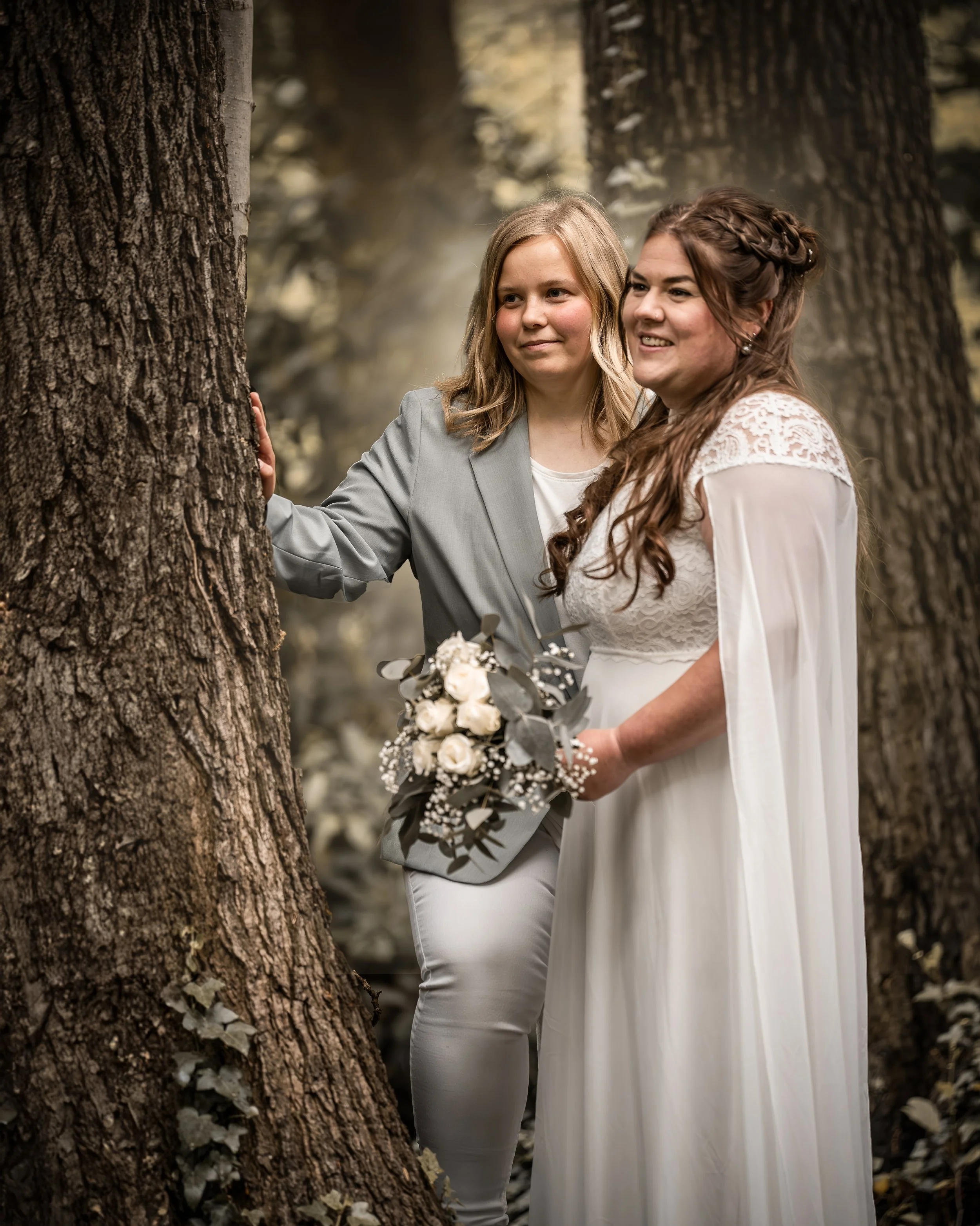 Zwei Frauen in Hochzeitskleidung im Wald, eine hält einen Brautstrauß, sie lächeln und stehen hinter einem Baum.