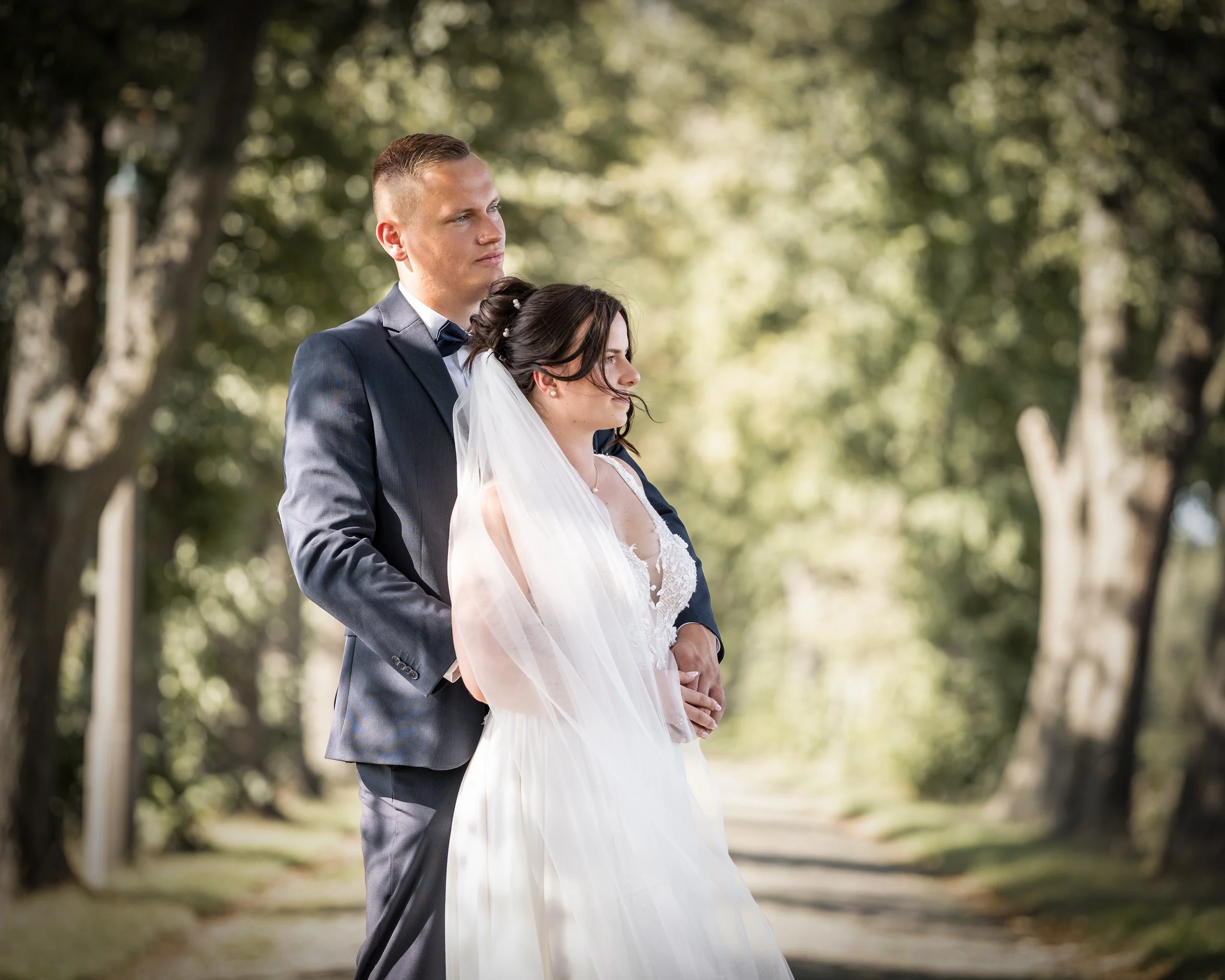 Ein Brautpaar steht im Freien unter Bäumen bei einer Hochzeit, die Braut trägt ein weißes Kleid mit Schleier, der Bräutigam trägt einen dunklen Anzug mit Fliege.