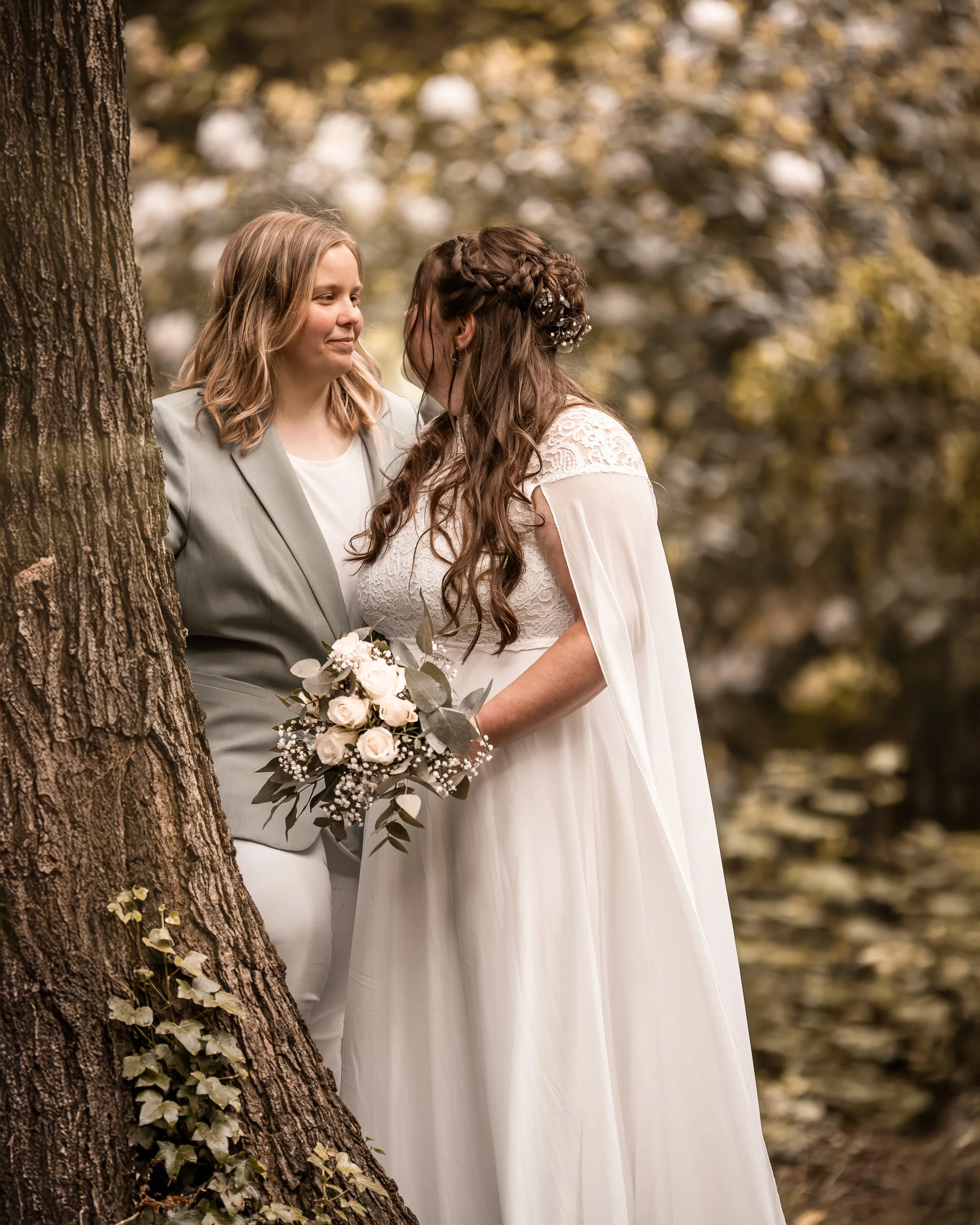 Zwei Frauen beim Blickkontakt im Wald, eine trägt ein weißes Brautkleid und die andere einen hellgrauen Anzug, um eine Hochzeit herum.