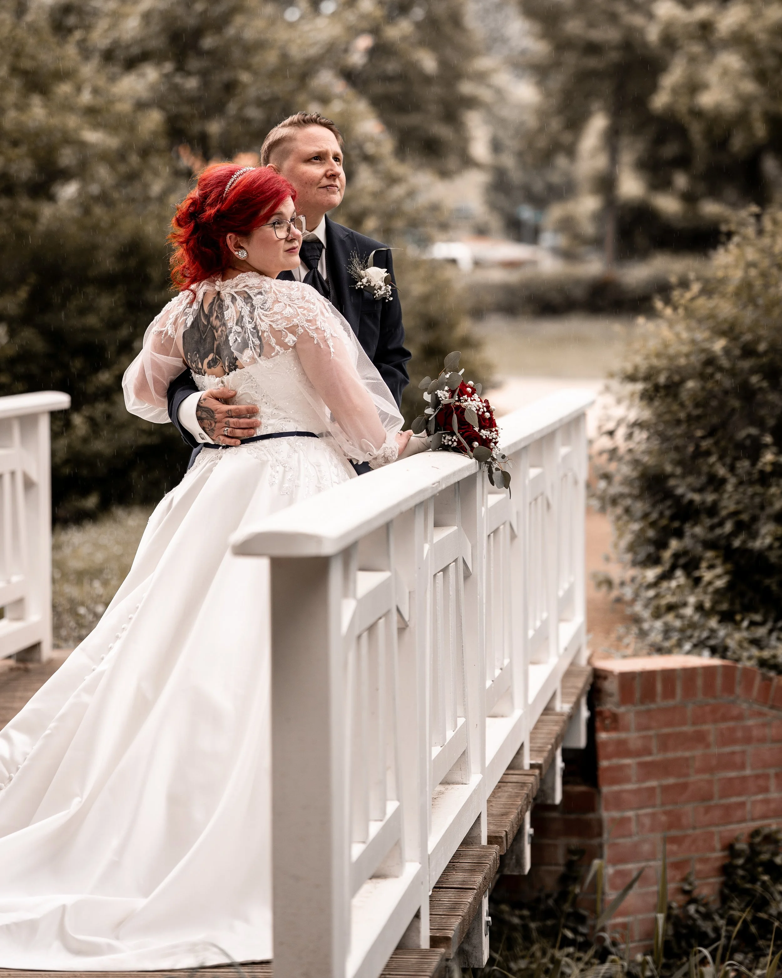 Paare stehen auf einer Brücke bei einer Hochzeit, mit Bäumen im Hintergrund und zuschauendem Publikum, bei Regen, während sie einander anschauen.