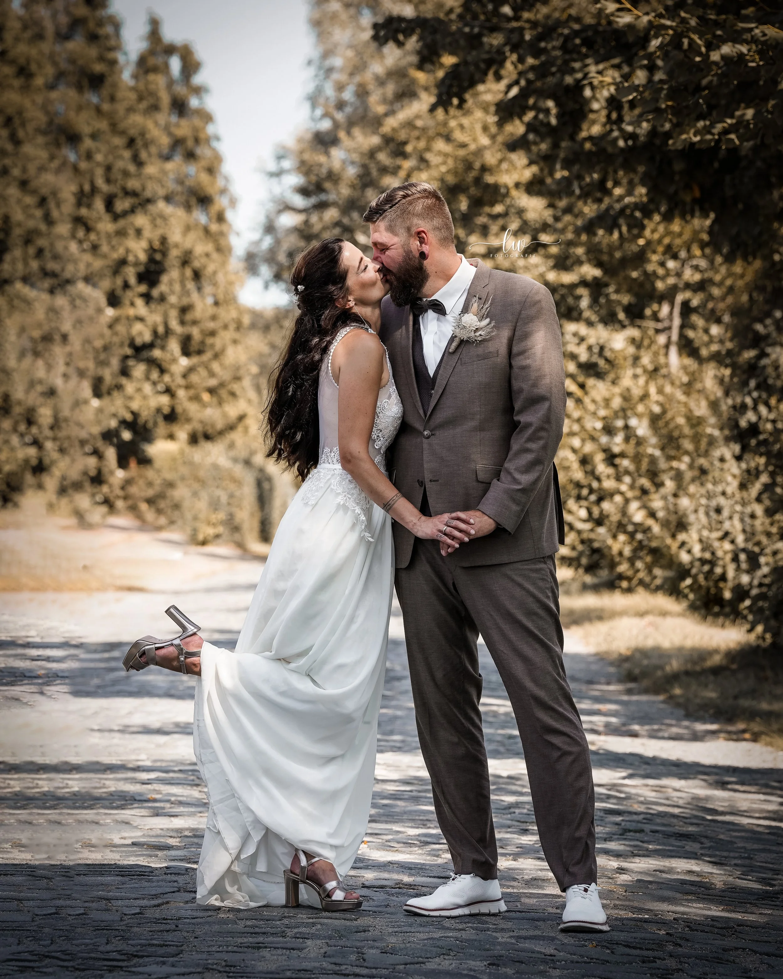 Ein Paar bei einer Hochzeit, das sich küsst, während sie auf einer gepflasterten Straße stehen, umgeben von Bäumen im Herbst.
