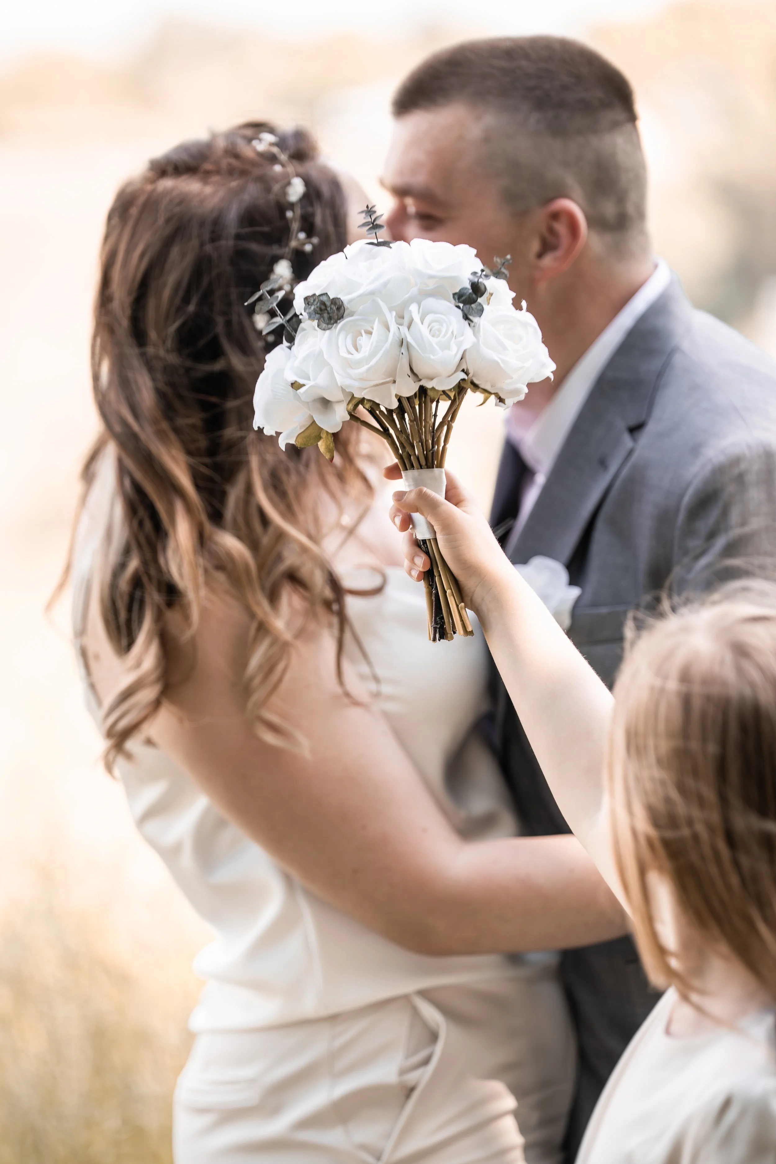 Ein Brautpaar bei einer Hochzeit, das sich küsst, während eine Person ein Blumenstrauß hält.