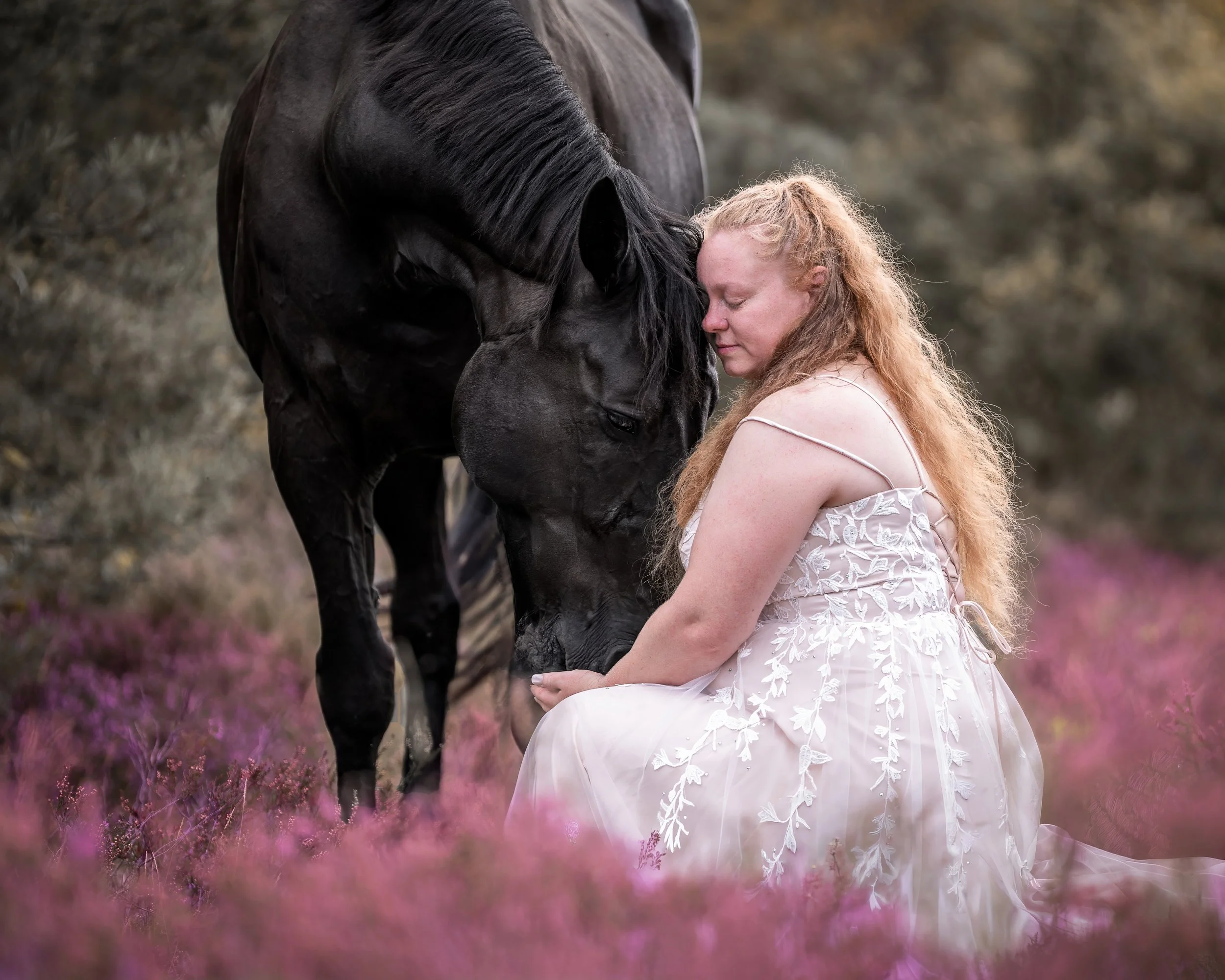 Eine rothaarige Frau in einem weißen, bestickten Kleid sitzt in einem Feld mit pinkfarbenen Blumen und ruht ihren Kopf auf die Stirn eines schwarzen Pferdes.