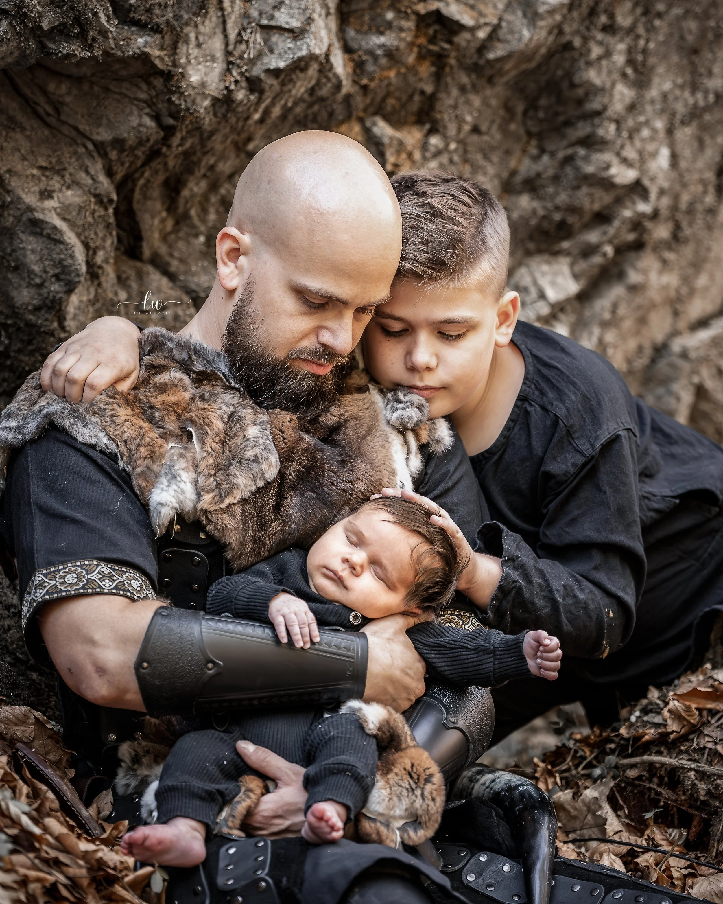 Ein Mann mit Glatze und Bart, ein Junge und ein Baby liegen auf dem Boden, umgeben von Laub, mit Tieren, die wie kleine Raubkatzen aussehen, alle sind in einer engen Umarmung und scheinen einen Moment der Zuneigung zu teilen.