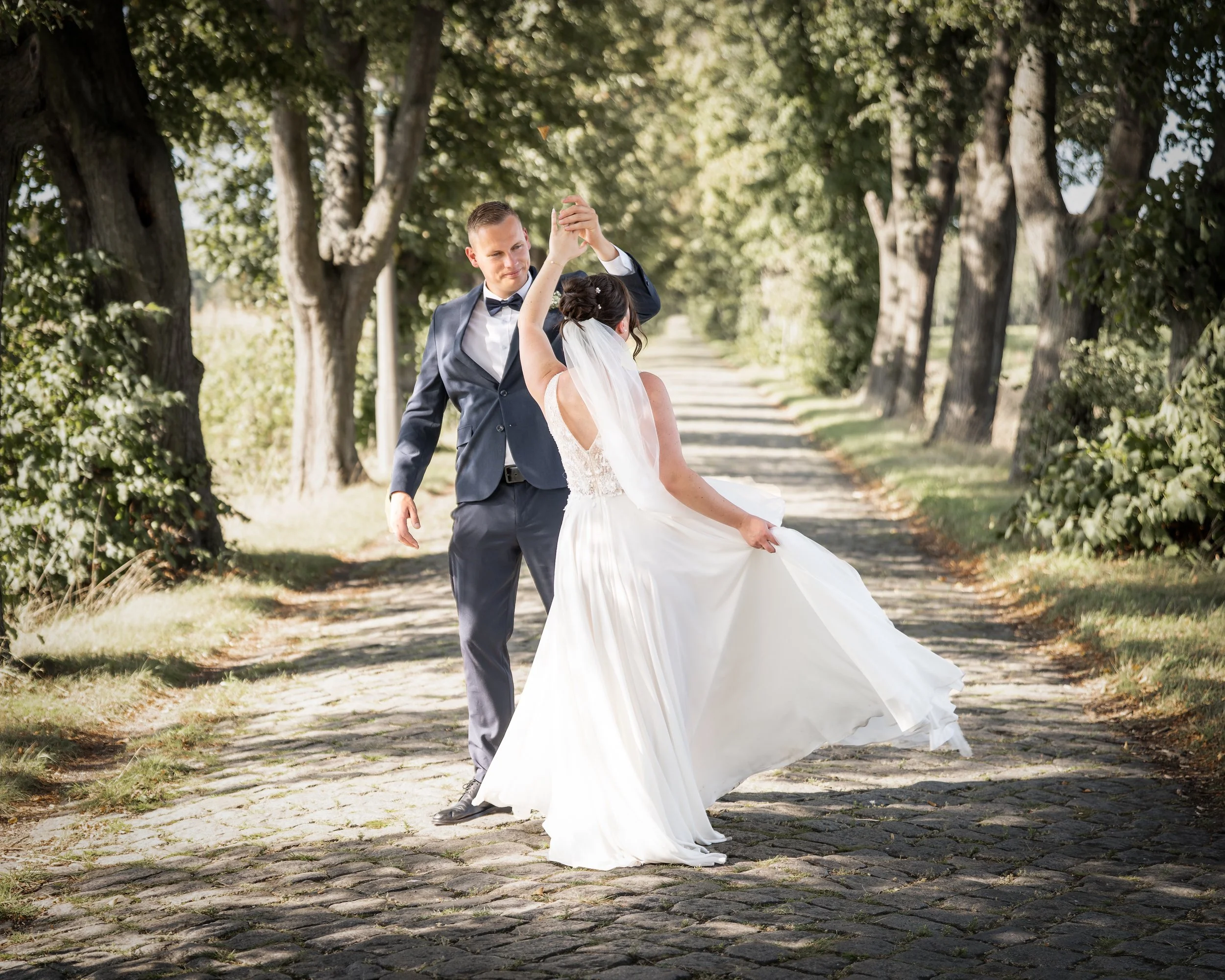 Ein Hochzeitsdarstellerpaar tanzt auf einem schmalen, gepflasterten Waldweg, umgeben von dichten Bäumen und Sonnenlicht, während die Braut in einem weißen Kleid und Schleier und der Bräutigam in einem schicken Anzug mit Fliege trägt.