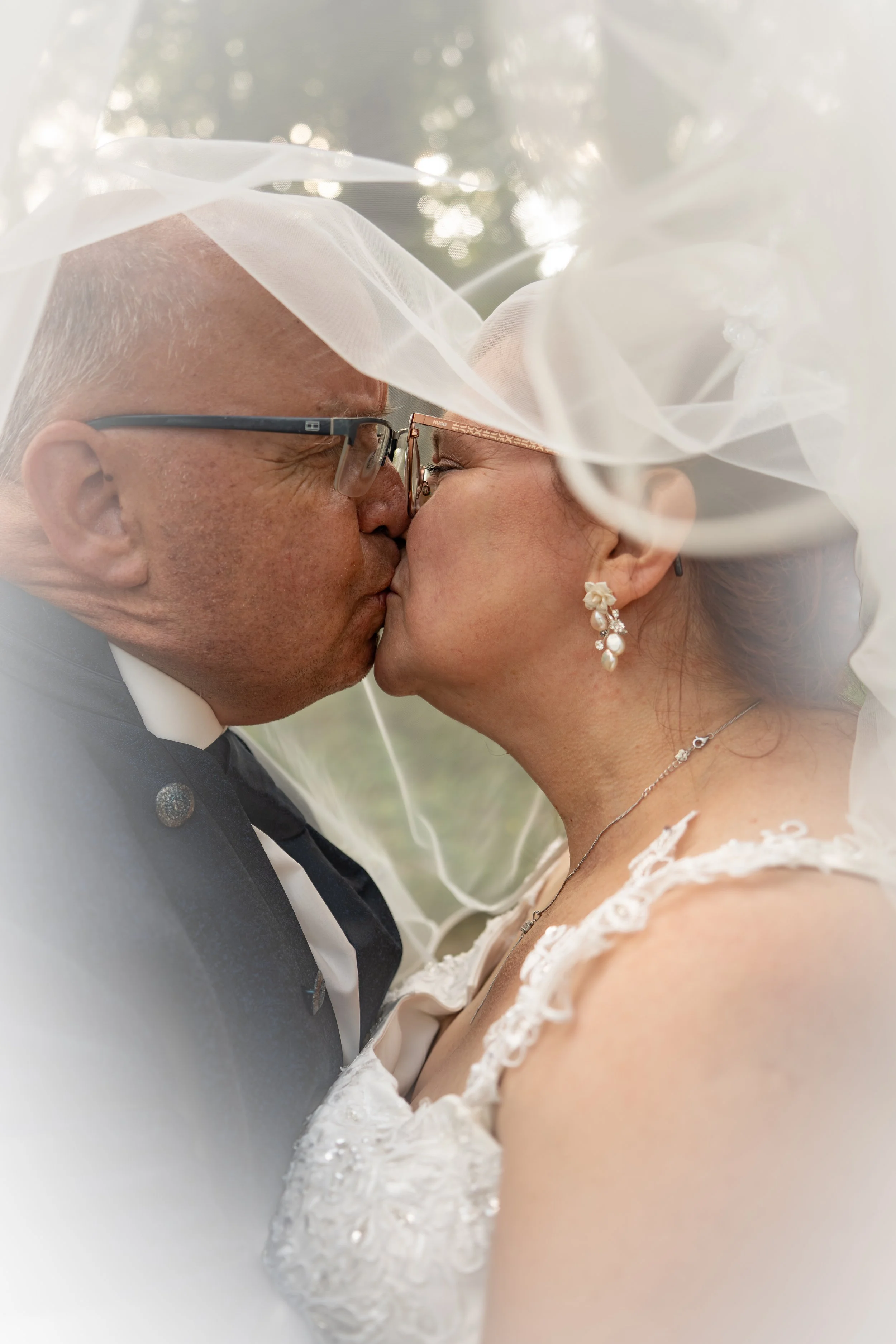 Ein frisch vermähltes Ehepaar teilt sich einen Kuss im Freien, die Frau trägt eine Hochzeitskleid und Ohrringe, der Mann trägt einen schwarzen Anzug. Der Hintergrund ist unscharf mit Lichtreflexen und Bäumen.