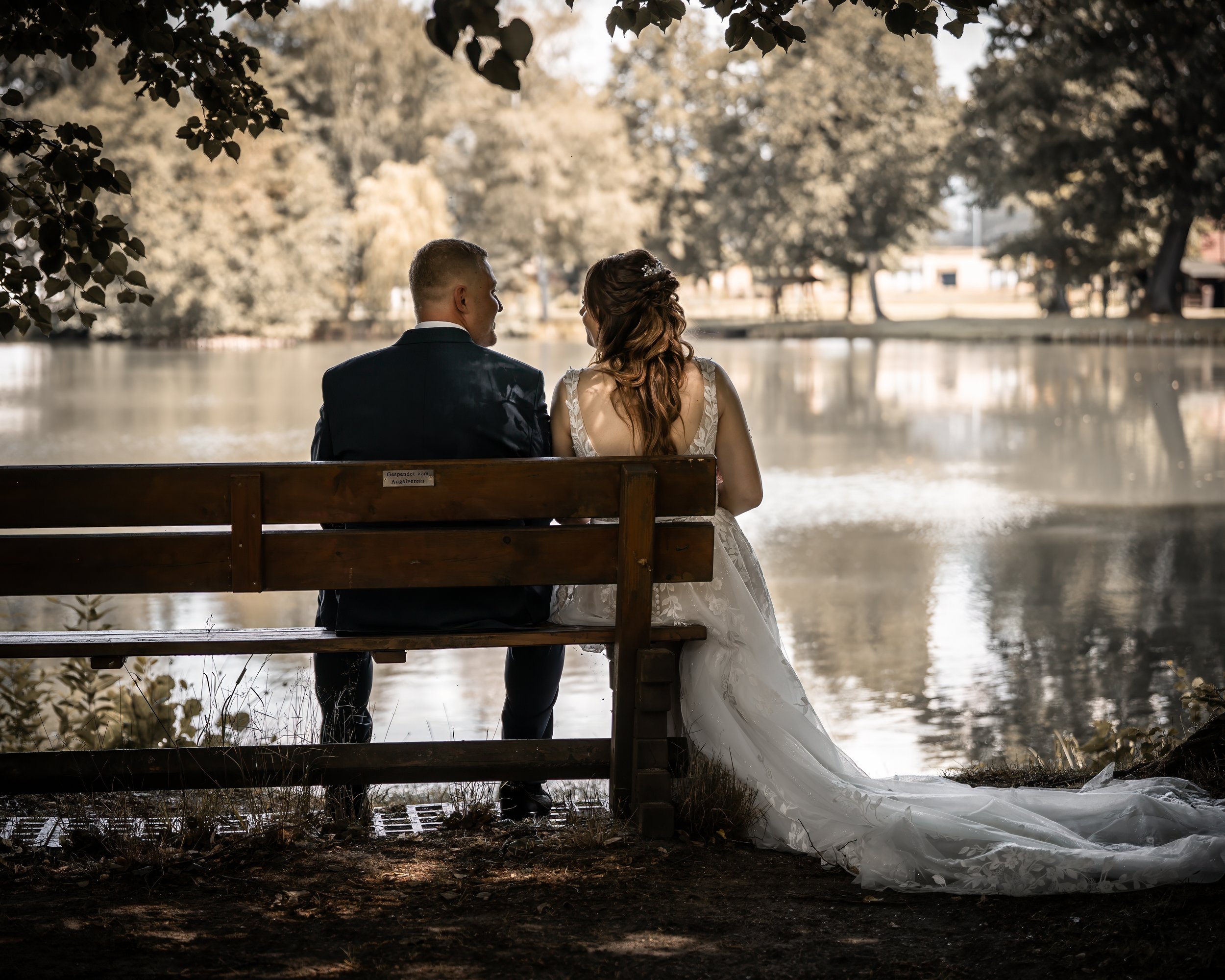 Ein Brautpaar sitzt auf einer Bank an einem See, die Braut trägt ein weißes Hochzeitskleid und der Bräutigam trägt einen dunklen Anzug, umgeben von Bäumen.