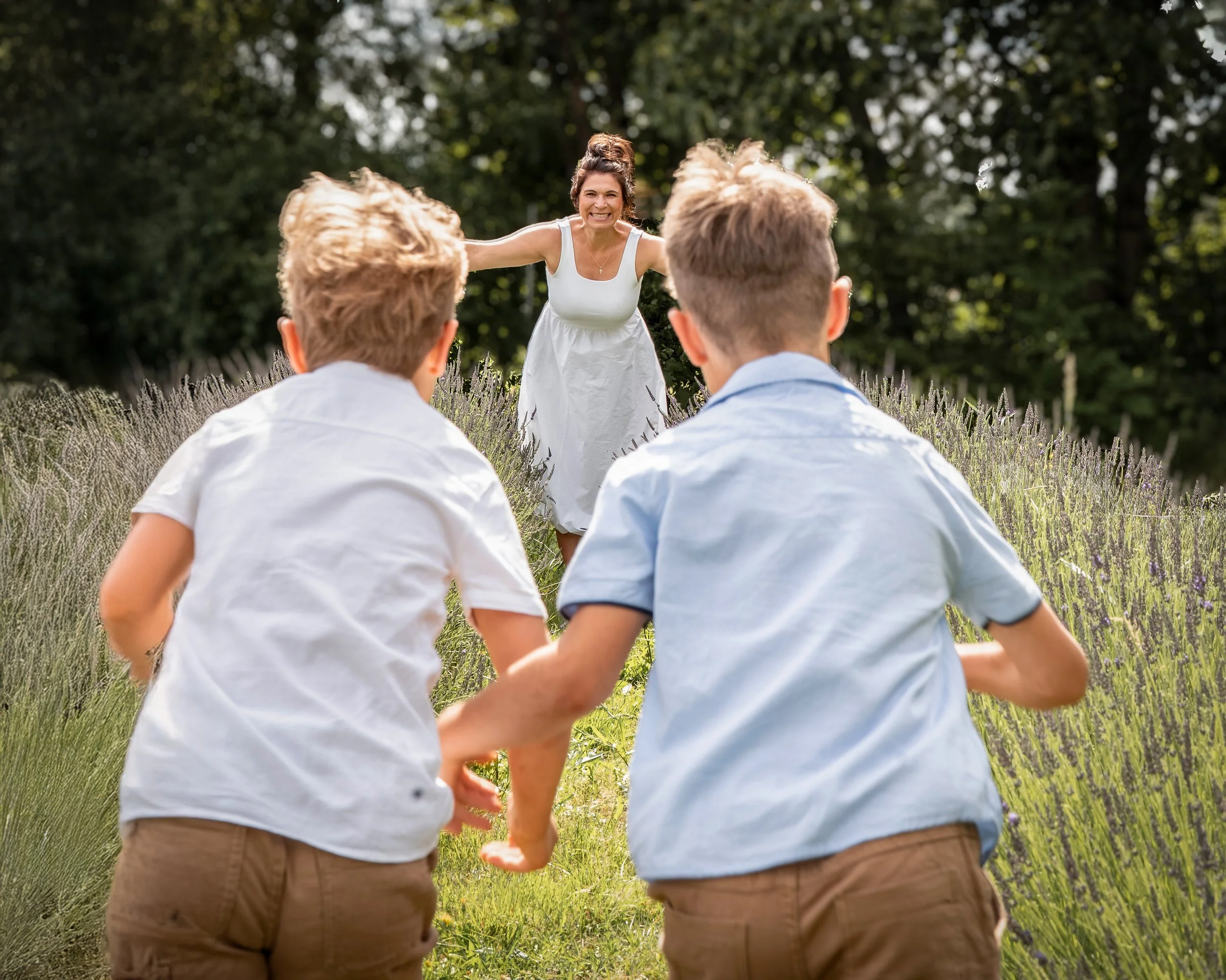 Eine Frau in einem weißen Kleid läuft Hand in Hand mit zwei Jungen in einem Lavendelfeld im Freien, umgeben von Bäumen, an einem sonnigen Tag.