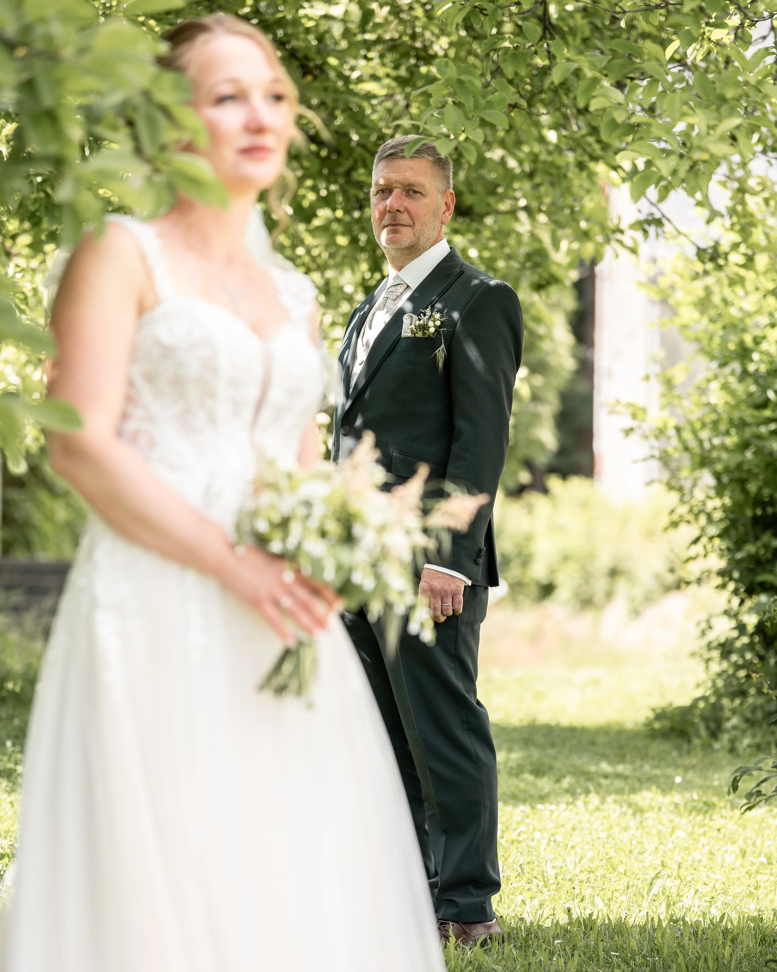 Ein Mann in einem schwarzen Anzug und eine Frau in einem weißen Brautkleid stehen im Garten unter einem Baum, vermutlich bei einer Hochzeitsfeier.