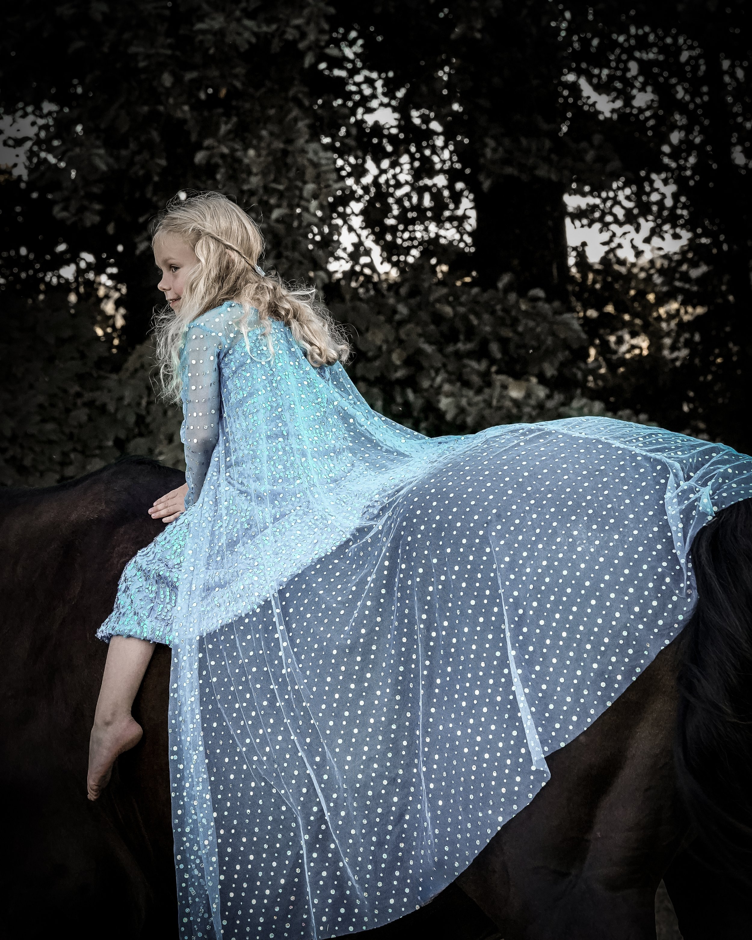 Ein Mädchen mit blonden, lockigen Haaren sitzt auf einer schwarzen Pferd; sie trägt ein langes, blaues Kleid mit weißen Punkten, das an den Schultern gerafft ist. Im Hintergrund stehen Bäume, nachts, Himmel sichtbar durch die Zweige.