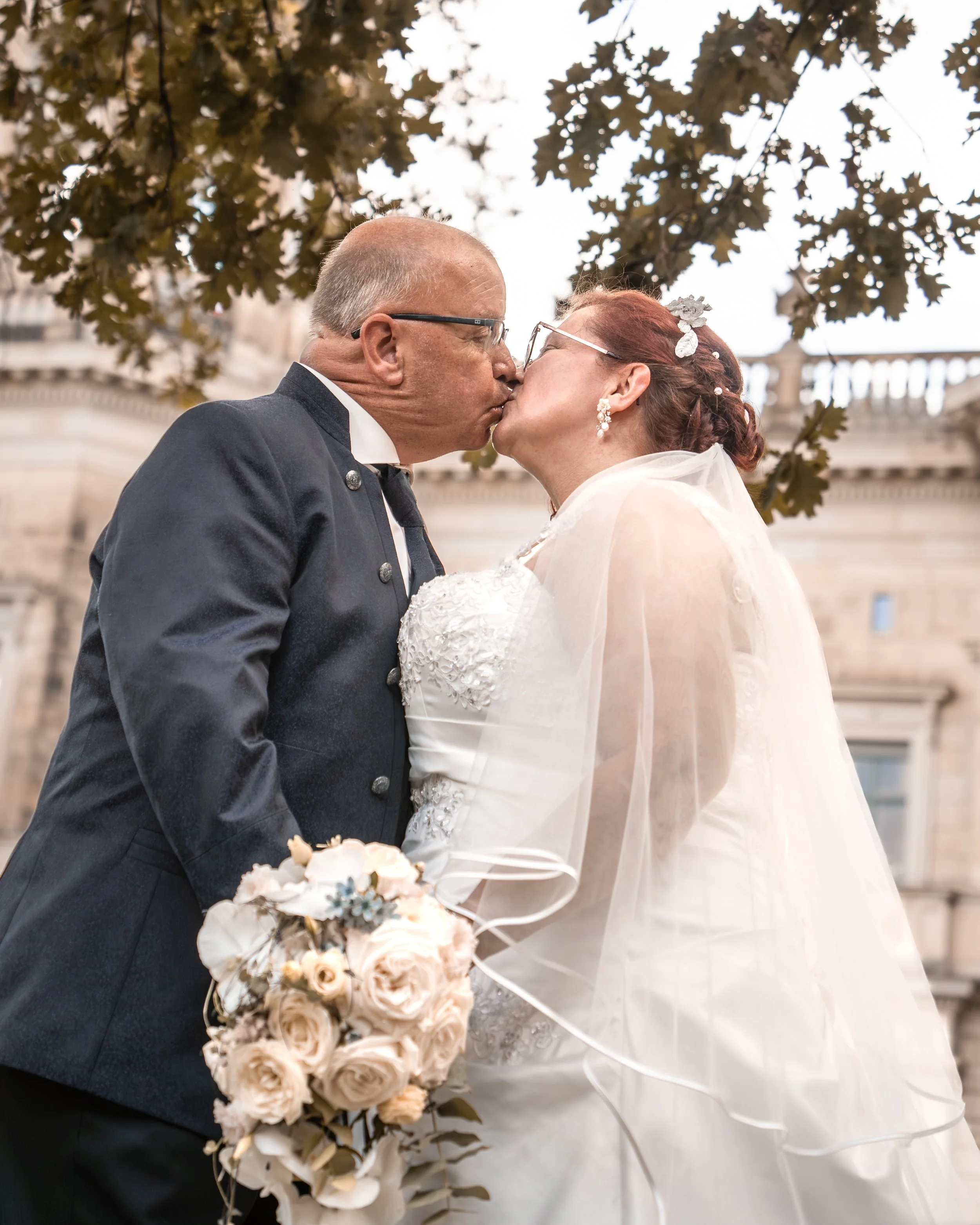 Ein älteres Paar bei einer Hochzeit, das sich küsst, während es unter einem Baum steht.