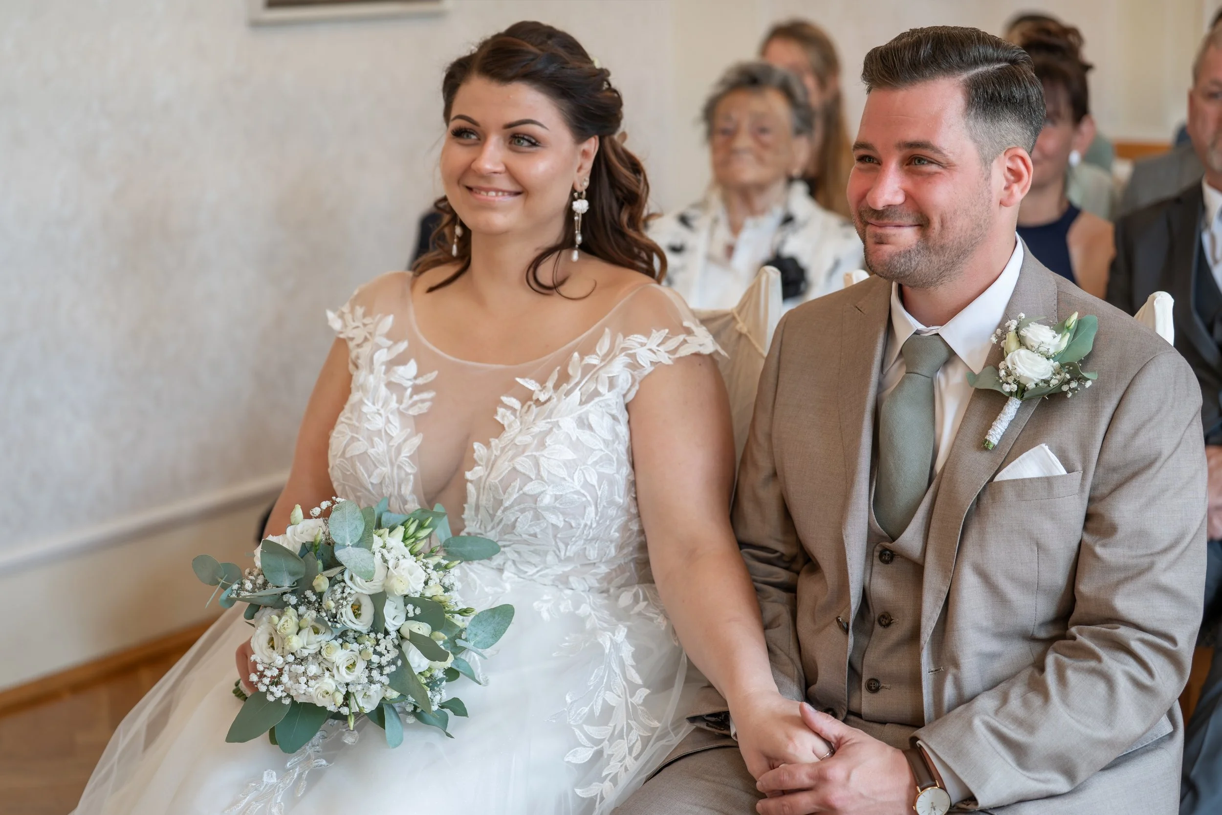 Braut und Bräutigam bei einer Hochzeit, sitzen nebeneinander, lächeln, die Braut hält einen Blumenstrauß, im Hintergrund Gäste.