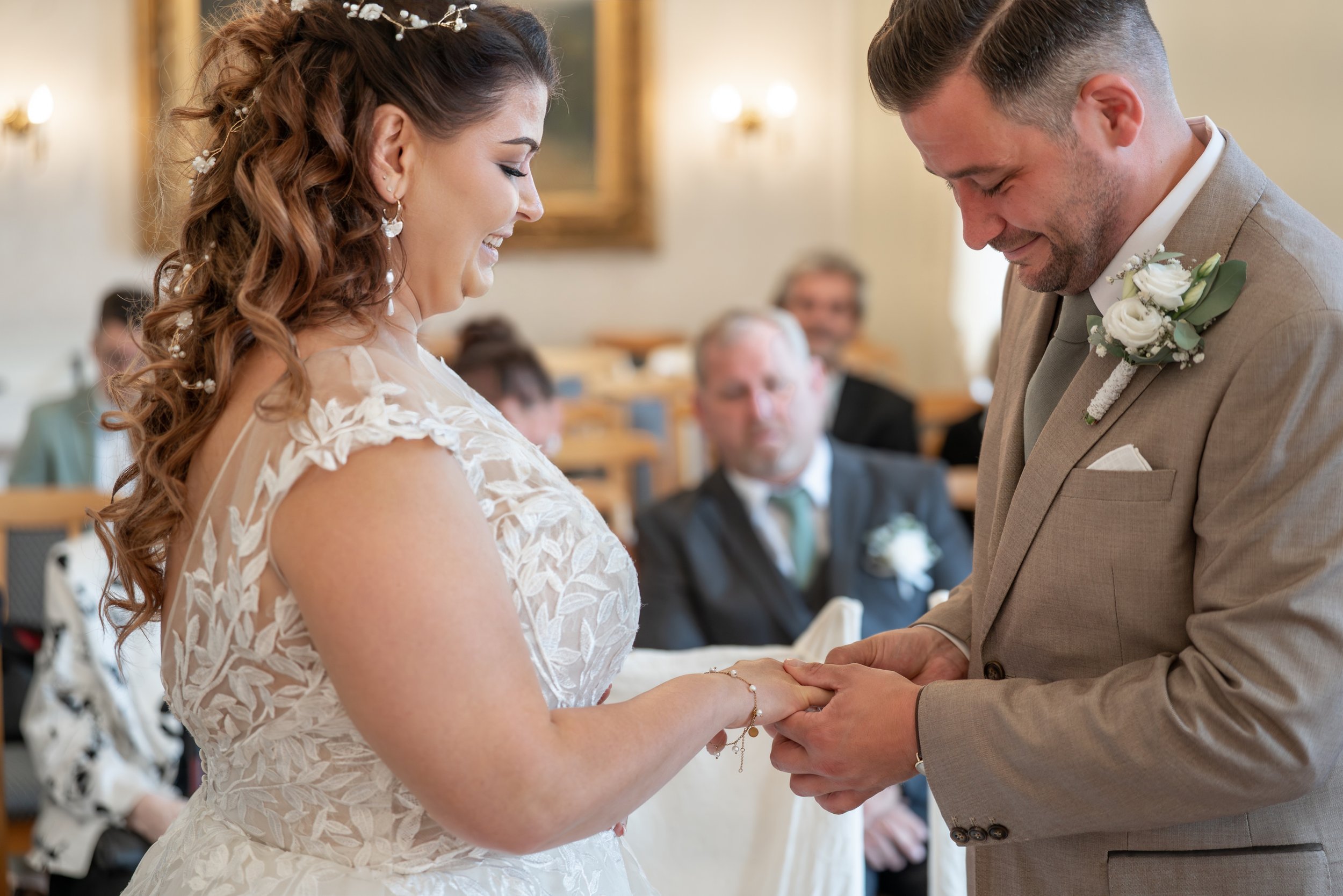 Braut und Bräutigam bei ihrer Hochzeit, halten sich an den Händen und schauen sich liebevoll an, im Hintergrund sind Hochzeitsgäste.