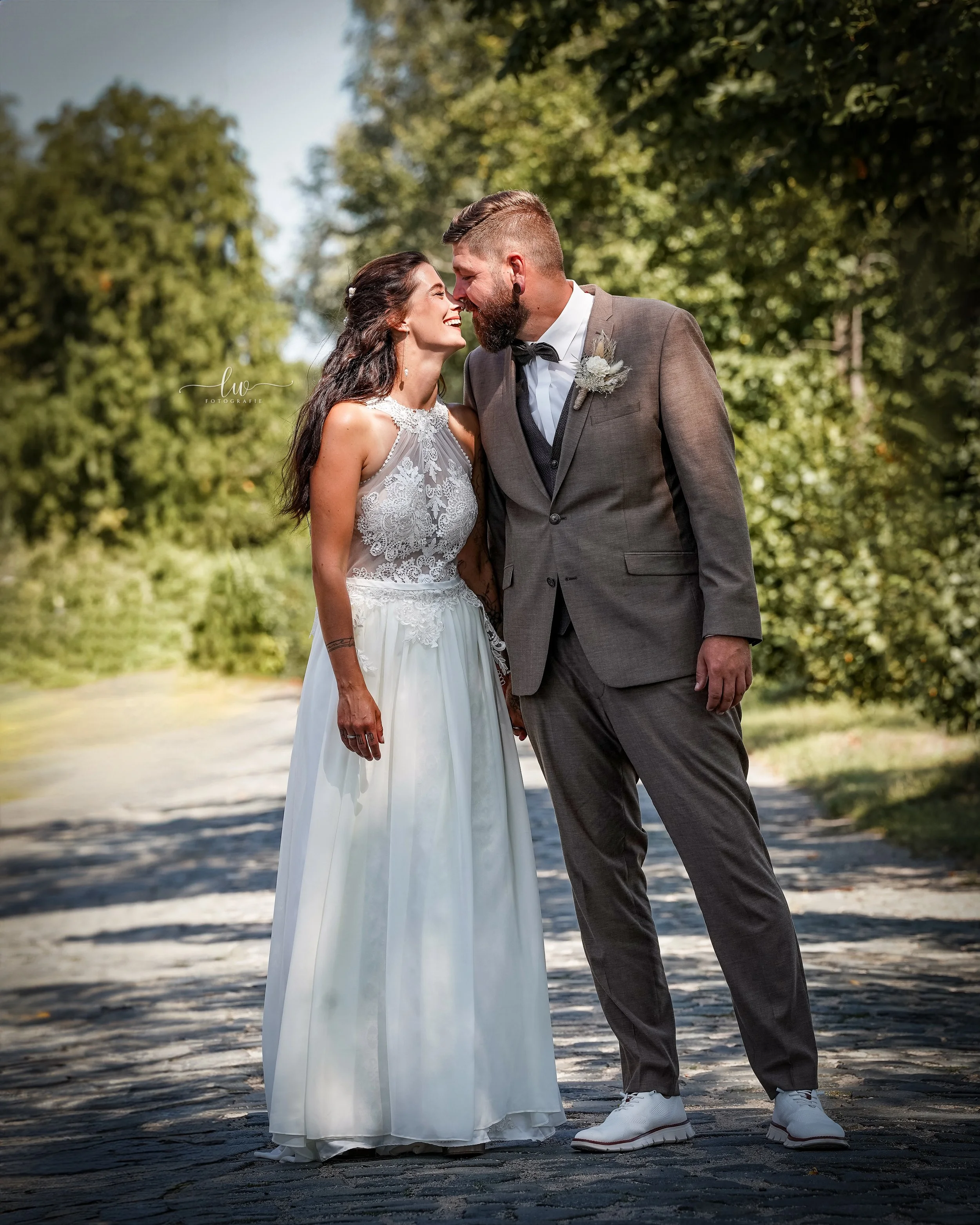 Ein Brautpaar, lachend und sich berührend, bei einer Hochzeit im Freien, umgeben von grünen Bäumen.