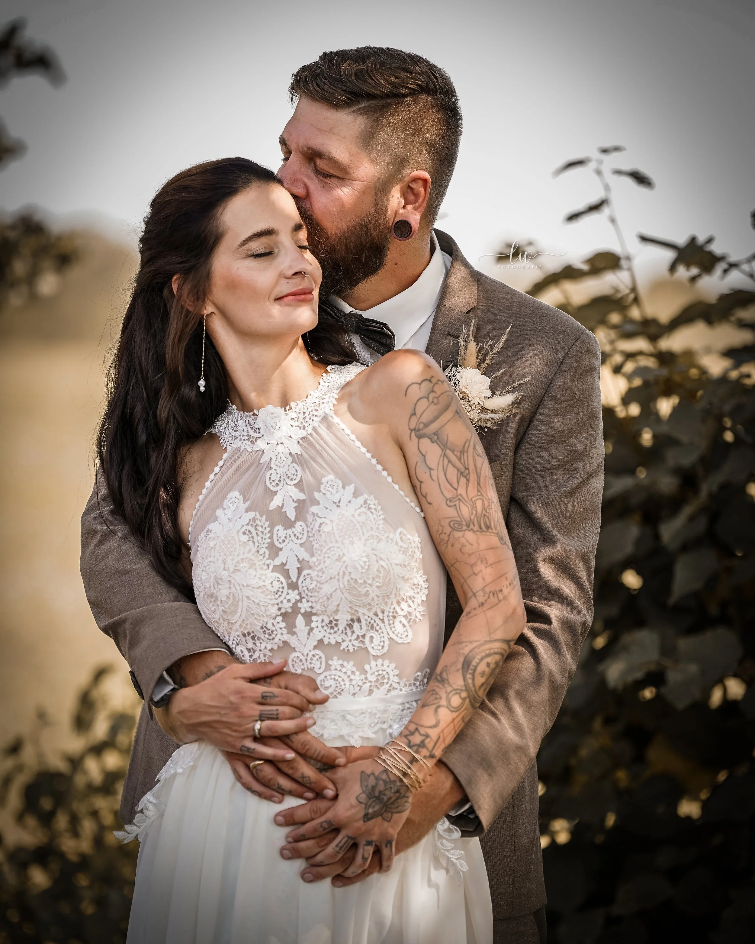 Ein Paar bei einer Hochzeit, umarmt sich im Freien bei Sonnenuntergang, die Frau trägt ein weißes Hochzeitskleid, er trägt einen grauen Anzug, beide lächeln entspannt.