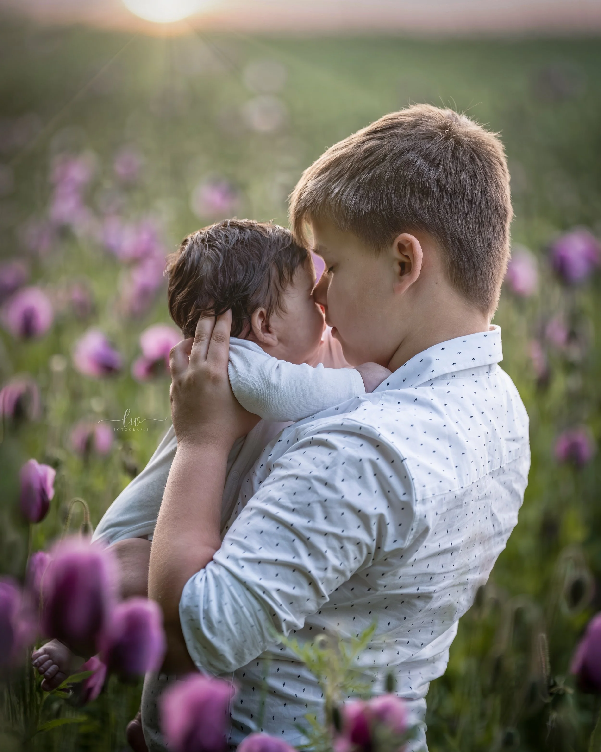Ein junger Mann hält ein Baby in einem Blumenfeld bei Sonnenuntergang. Die beiden teilen einen intimen Moment, die Sonne strahlt im Hintergrund.