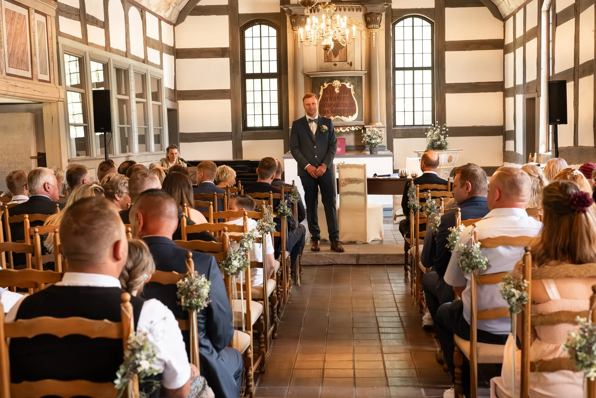Ein Mann in Anzug und Fliege steht vor einer Gruppe von Menschen in einem festlich dekorierten Raum, vermutlich bei einer Hochzeit.