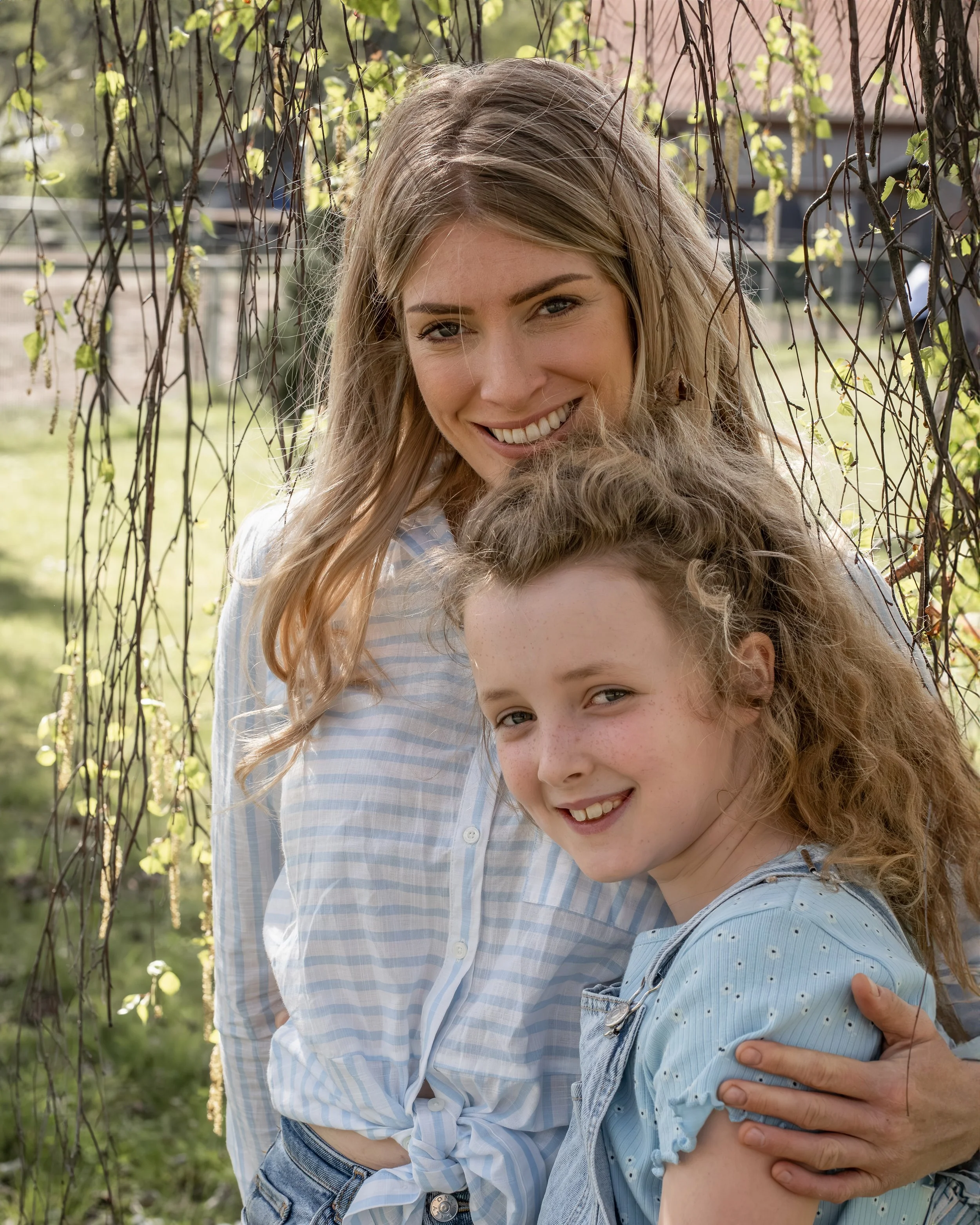 Zwei lachende Mädchen, eines mit langen blonden Haaren und das andere mit lockigen braunen Haaren, umarmend im Freien auf einer grünen Wiese, mit Baumzweigen im Hintergrund.