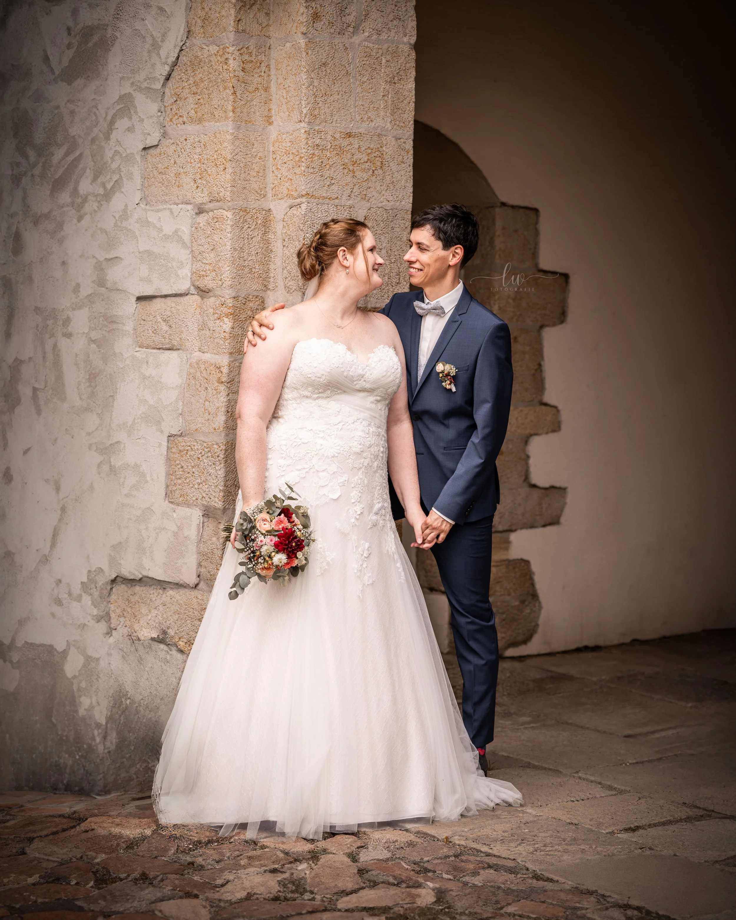 Ein Brautpaar hält Händchen und schaut sich verliebt an, während die Braut ein weißes Hochzeitskleid und der Bräutigam einen blauen Anzug trägt. Sie stehen vor einer alten Steinfassade.