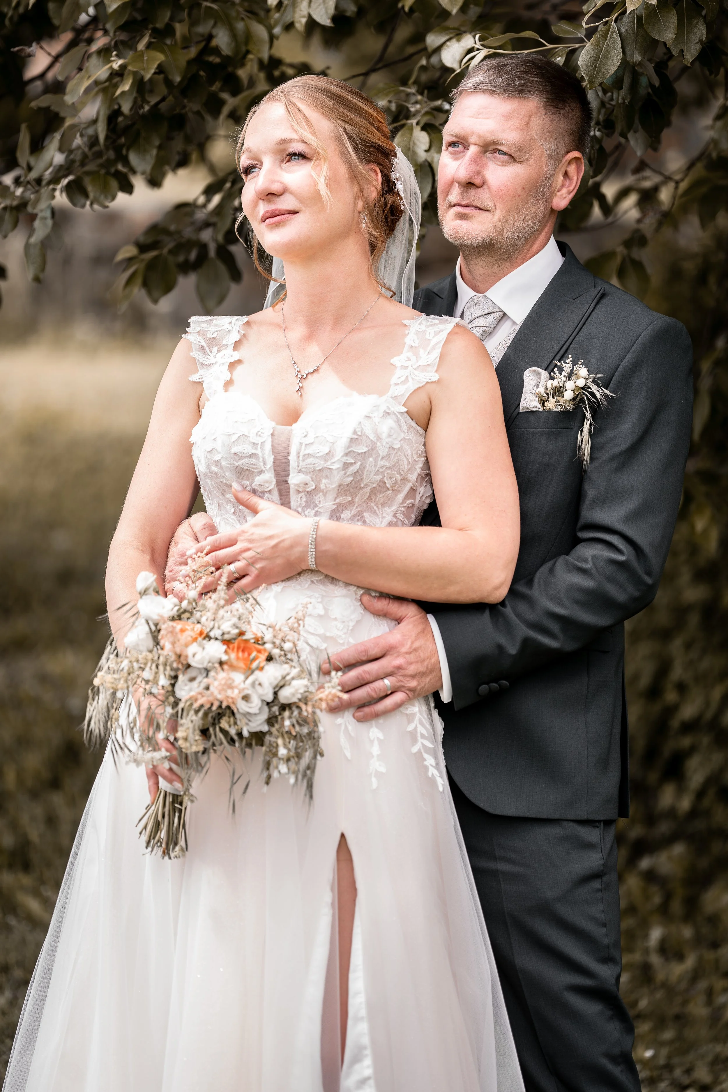 Ein Brautpaar steht draußen vor einem Baum. Die Braut trägt ein weißes Hochzeitskleid mit Spitze und eine silberne Halskette. Der Bräutigam trägt einen dunklen Anzug mit einer weißen Krawatte und hat eine Boutonnière am Revers. Sie halten sich an den Händen, die Braut hält einen Blumenstrauß.