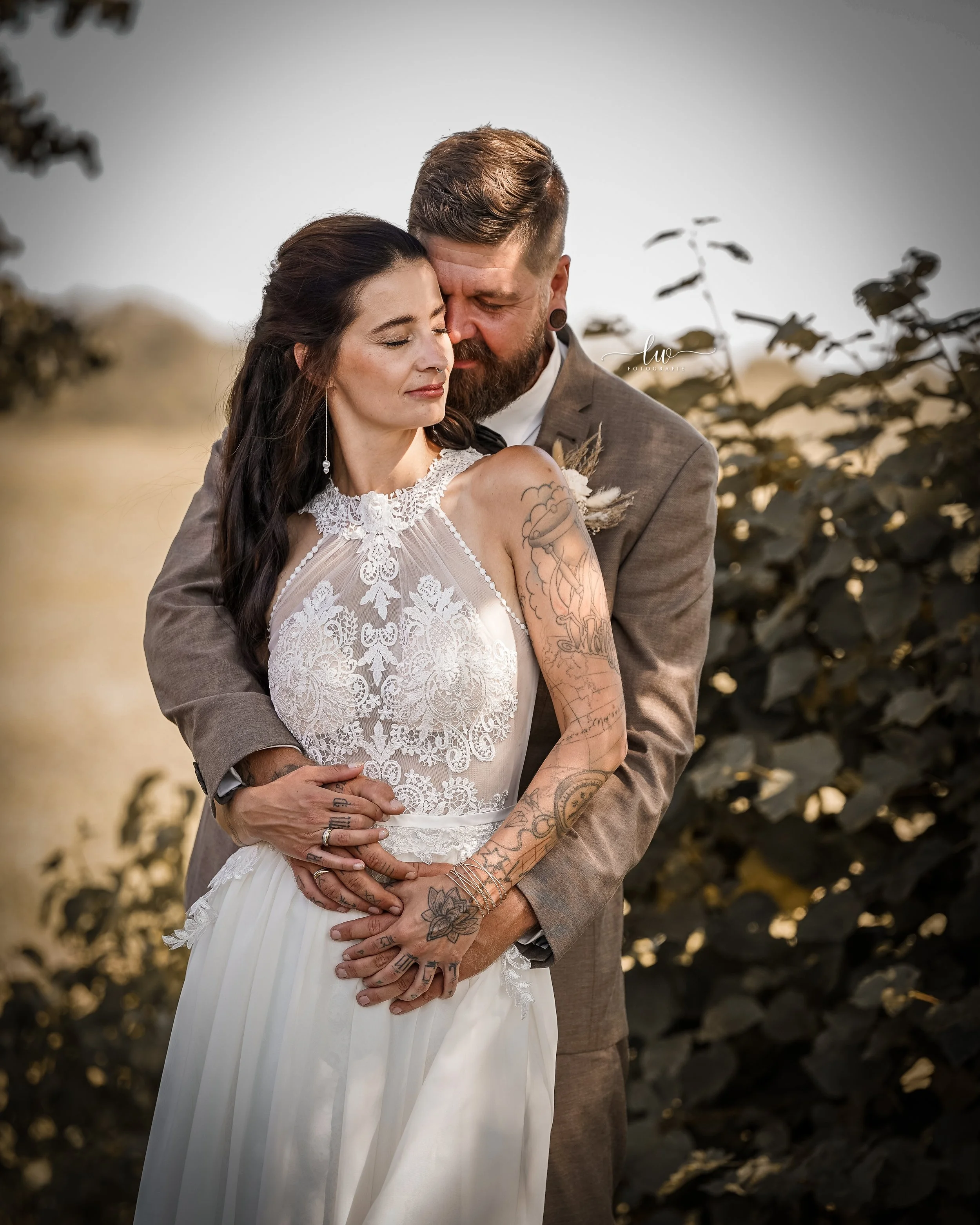 Ein verliebtes Brautpaar steht draußen umarmt, die Frau trägt ein weißes Hochzeitskleid mit Spitzen und Tattoos am Arm, der Mann trägt einen grauen Anzug mit Kurzhaarschnitt und Bart. Hintergrund ist unscharf mit dunklen Blättern.