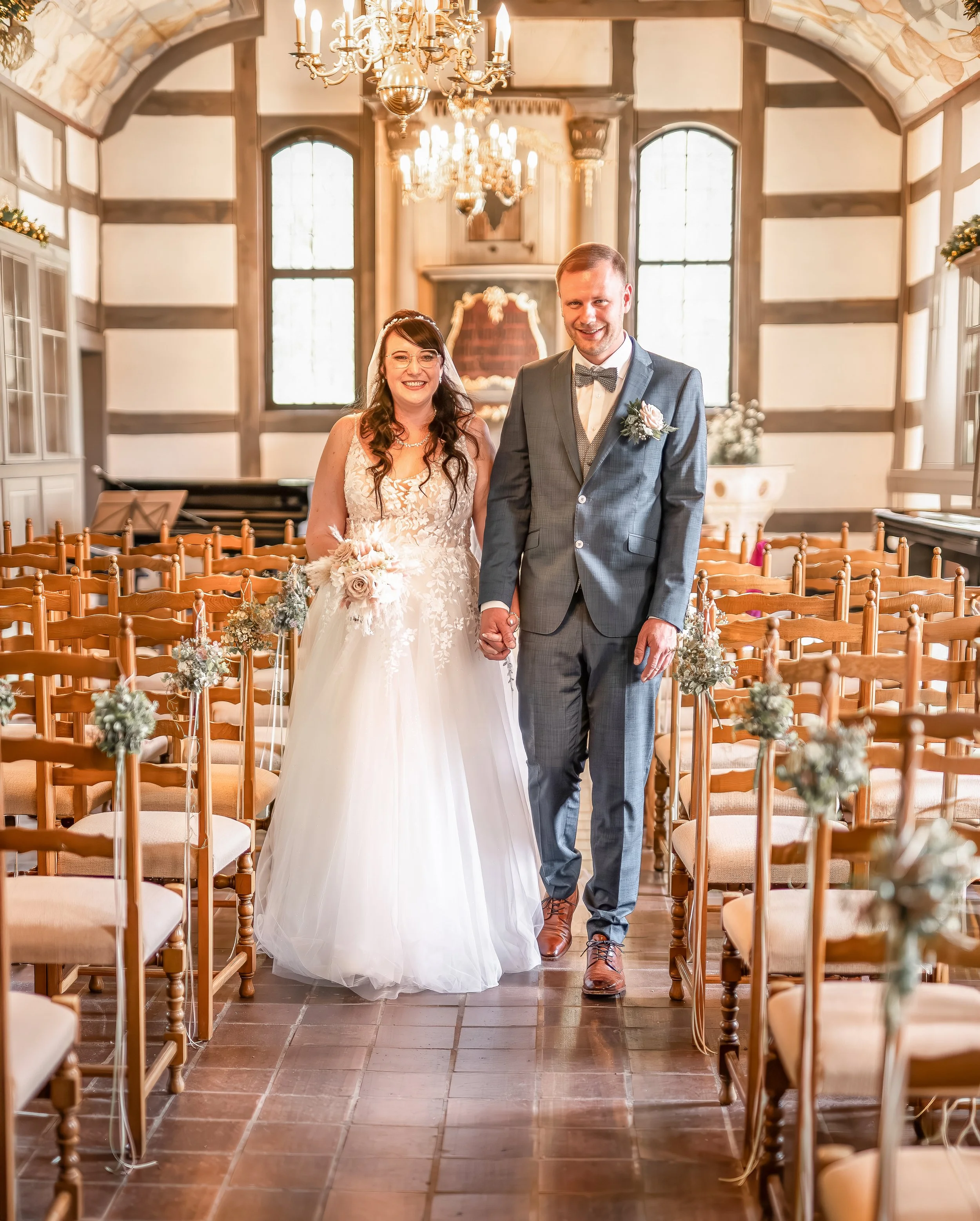 Ein Brautpaar steht in einer festlich dekorierten Kirche. Die Braut trägt ein weißes Hochzeitskleid mit Spitzenmuster und Halstuch, während der Bräutigam einen dunkelgrauen Anzug mit Fliege trägt. Beide halten sich an den Händen und lächeln. Der Raum ist mit Stühle im Vordergrund und einem Kronleuchter an der Decke ausgestattet.