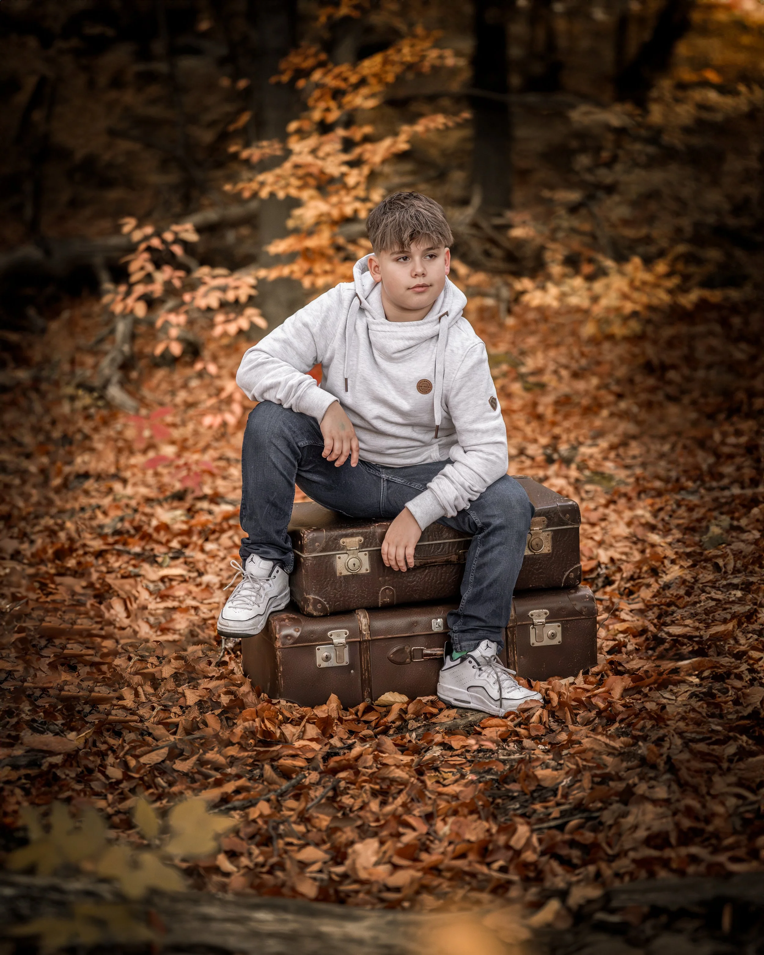 Junge auf einem Holzstuhl sitzt in einem herbstlichen Laubwald, trägt einen grauen Hoodie und dunkle Hosen.