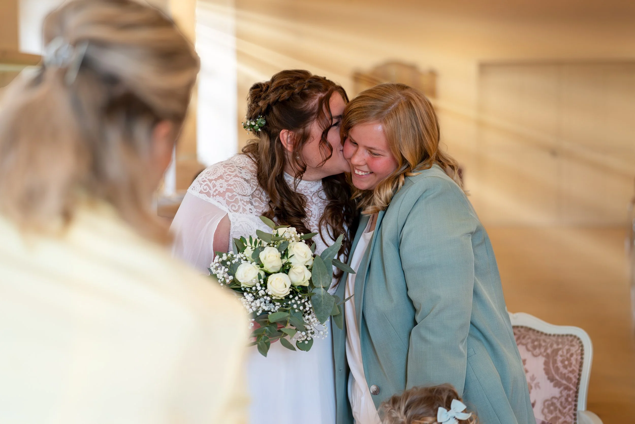 Zwei Frauen bei einer Hochzeit, eine küsst die andere auf die Wange, beide lächeln glücklich. Im Vordergrund eine weitere Person ist leicht unscharf zu erkennen.
