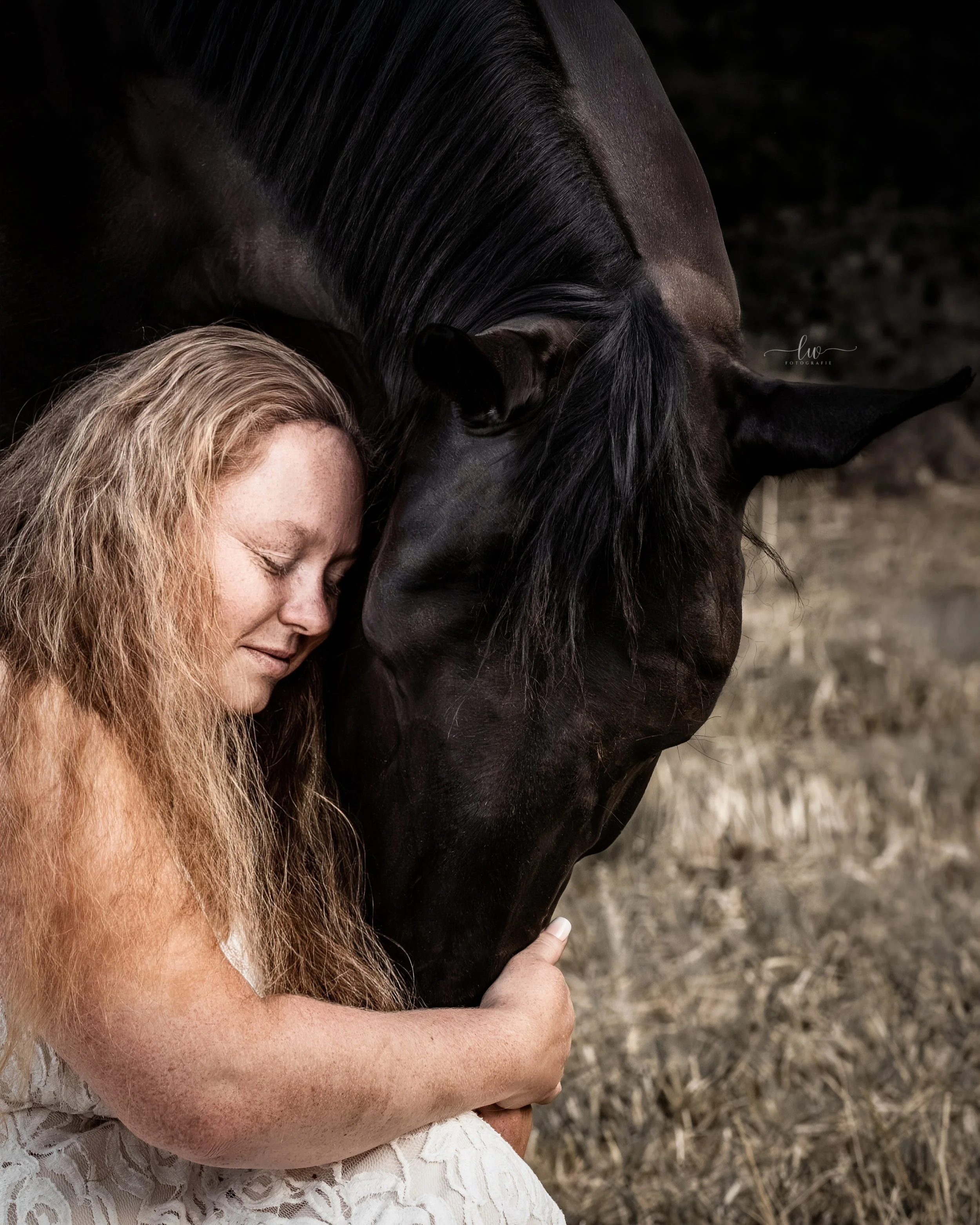 Frau mit lockigem, blondem Haar umarmt schwarzen Pferd, beide schließen die Augen, im Hintergrund eine Wiese mit trockenem Gras.