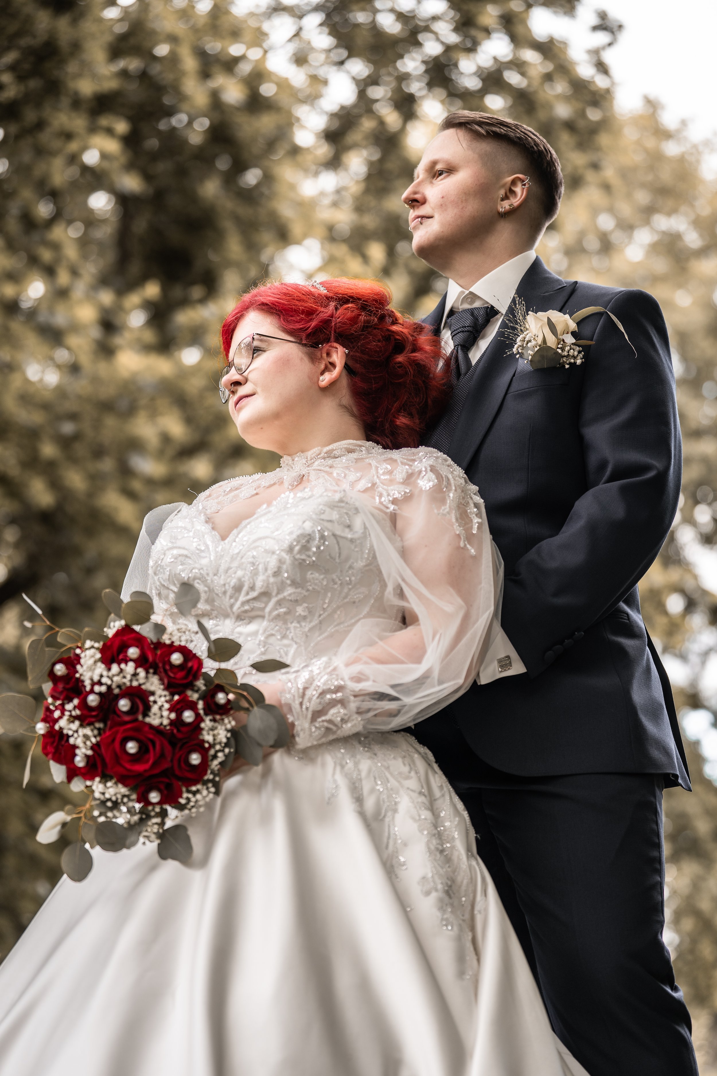 Ein Brautpaar bei einer Hochzeitsfotografie im Freien, mit dem Bräutigam in einem dunklen Anzug und der Braut in einem weißen Brautkleid mit roten Rosen im Brautstrauß.
