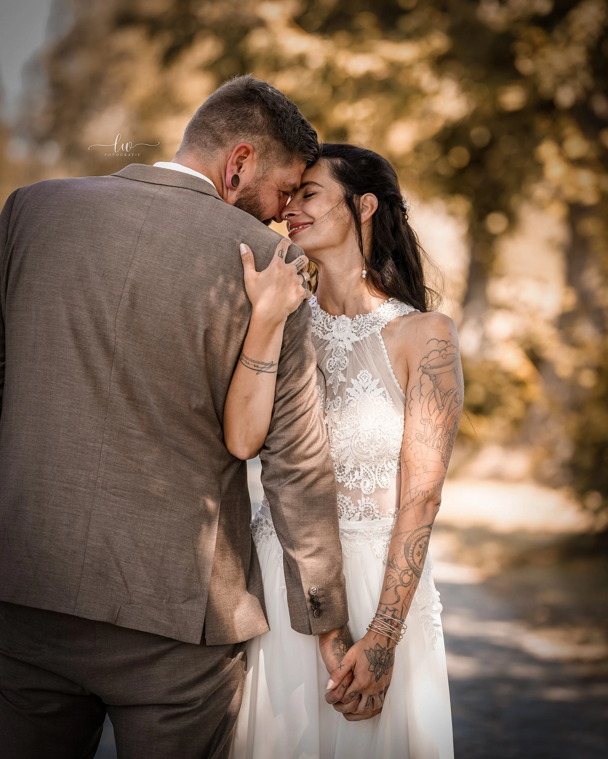 Ein Paar bei einer romantischen Umarmung im Freien bei Sonnenuntergang. Der Mann trägt einen grauen Anzug und die Frau ein weißes Brautkleid mit Tattoos an den Armen. Sie halten Händchen und lächeln sich liebevoll an.