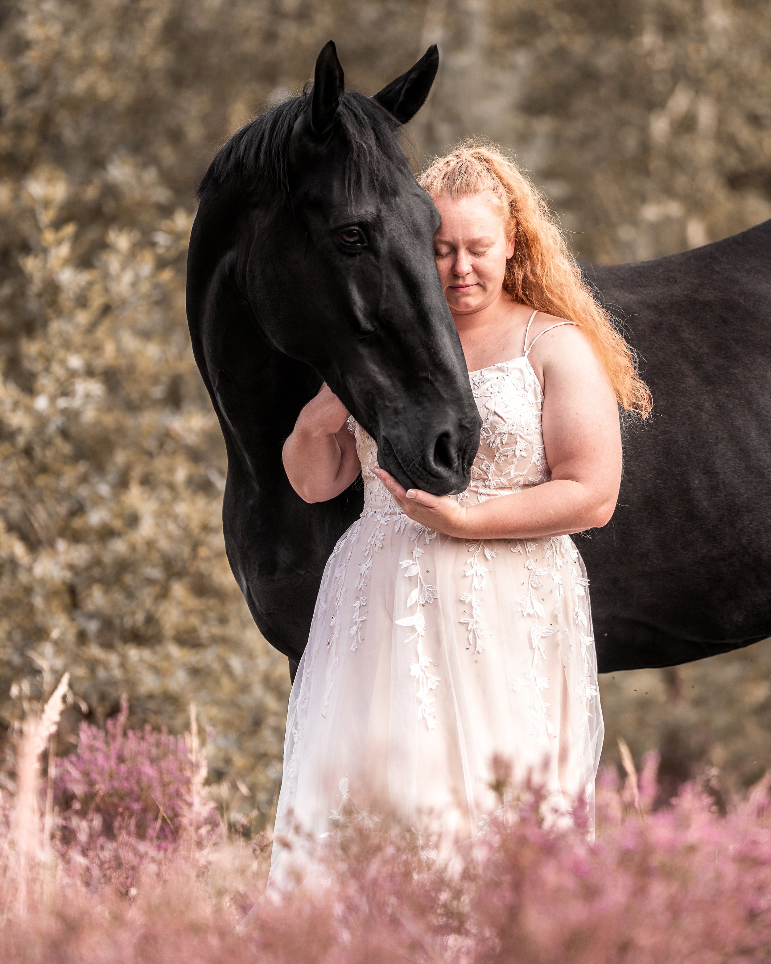 Eine Frau in einem weißen, eleganten Kleid steht mit einem schwarzen Pferd in einem natürlichen Außenbereich mit Blüten und Bäumen.