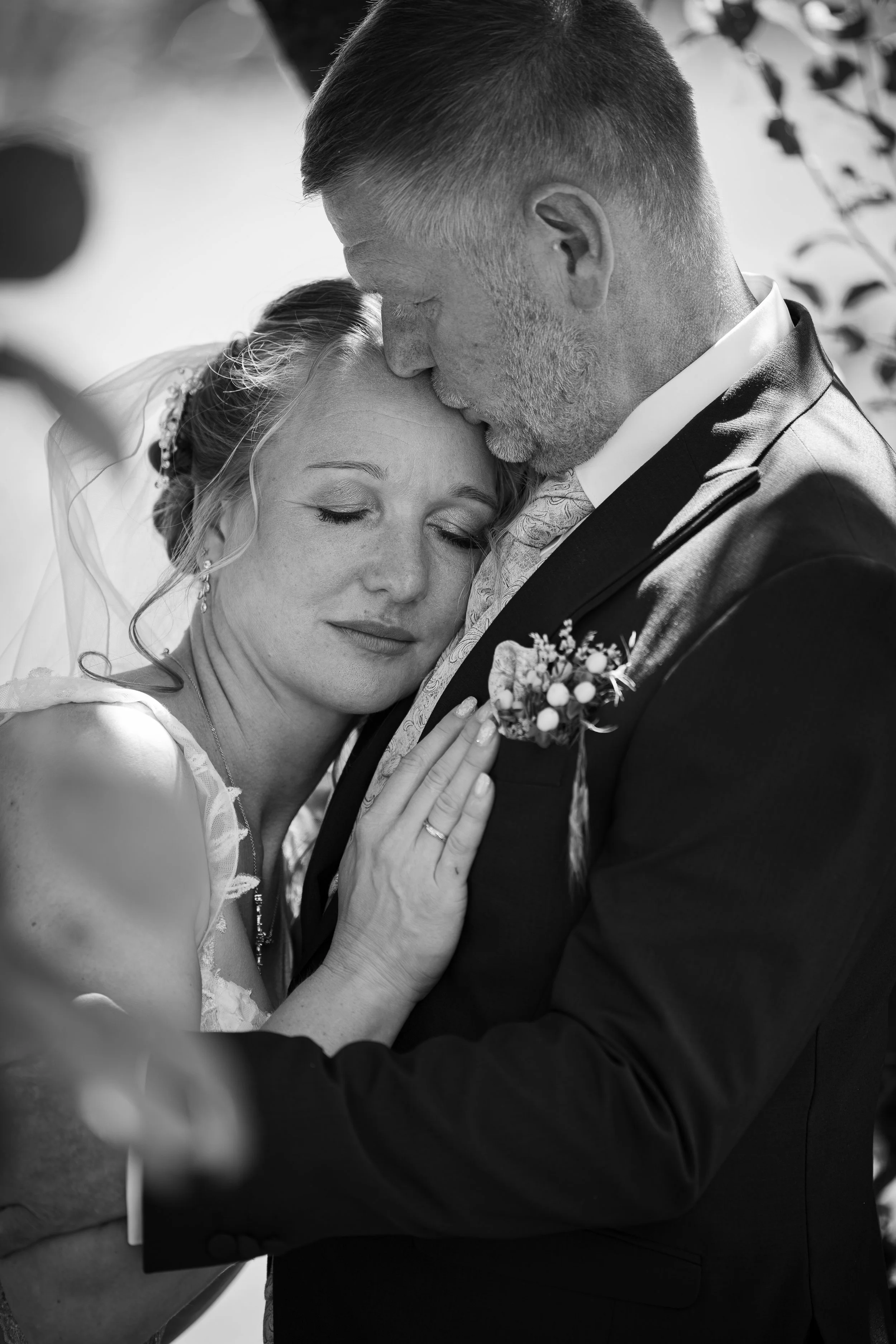 Ein Mann und eine Frau, die bei einer Hochzeit emotionalen Moment teilen, umarmt sich innig, während der Mann die Stirn der Frau küsst, beide in formaler Kleidung, schwarz-weiß Foto.