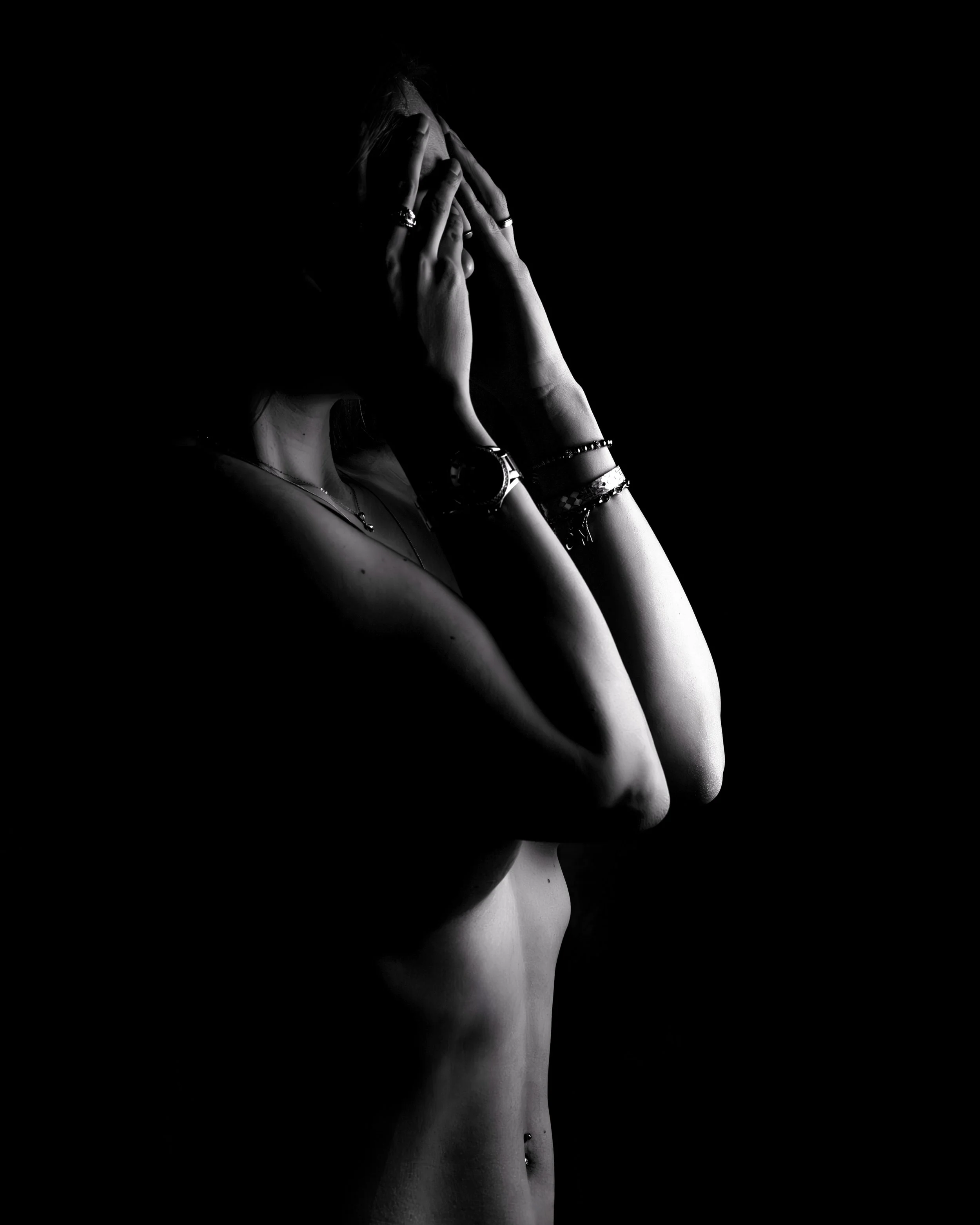 Schwarzes und weißes künstlerisches Portrait einer nackten Frau im Profil, Hände vor das Gesicht, dunkler Hintergrund, Fokus auf Gesicht und Arme, Schmuck an Händen und Arm, zartes sichtbar.