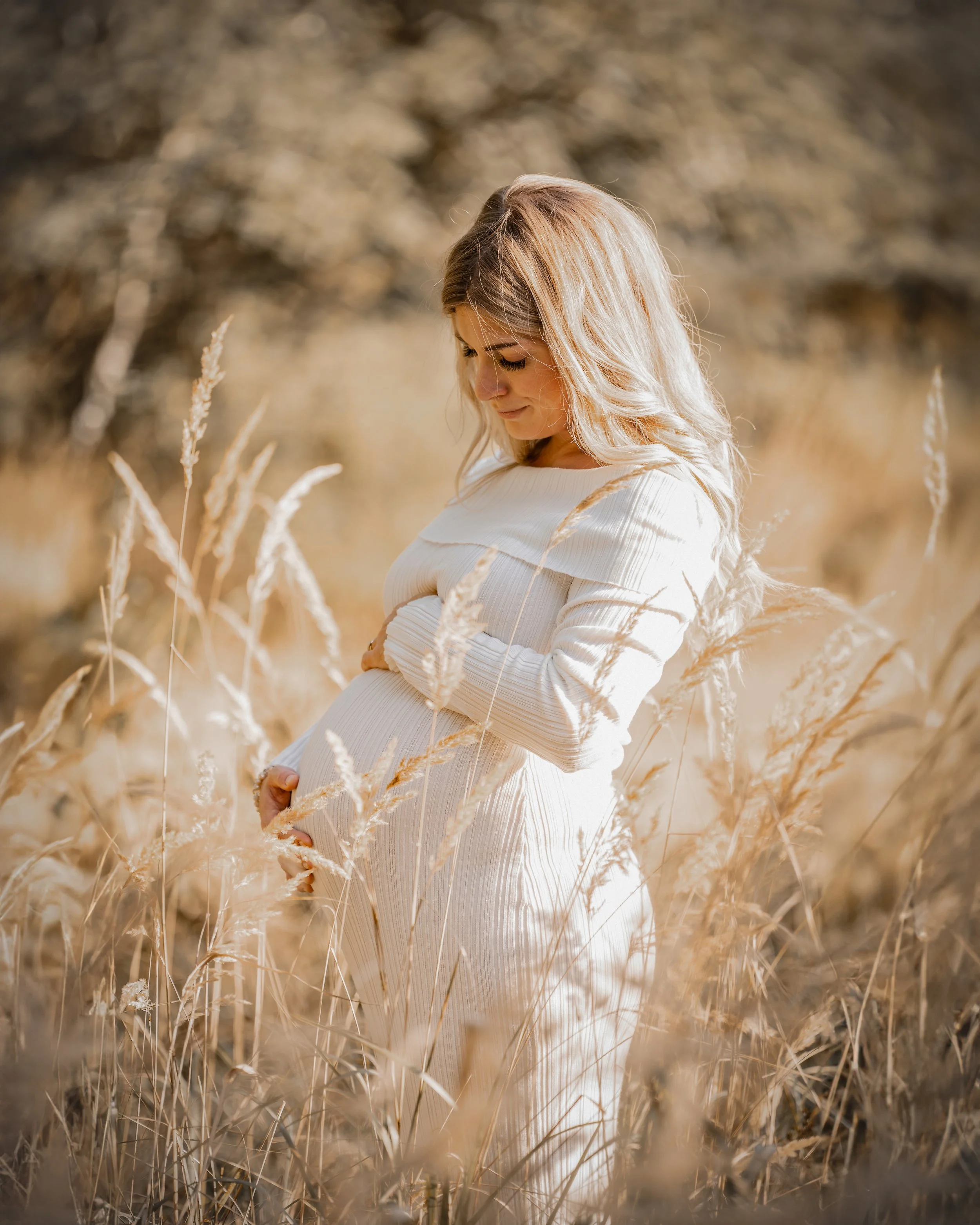 Eine schwangere Frau steht in einem goldenen Feld von Gräsern, trägt ein weißes Kleid und schaut nach unten, während sie ihre Hände auf ihren Bauch legt.