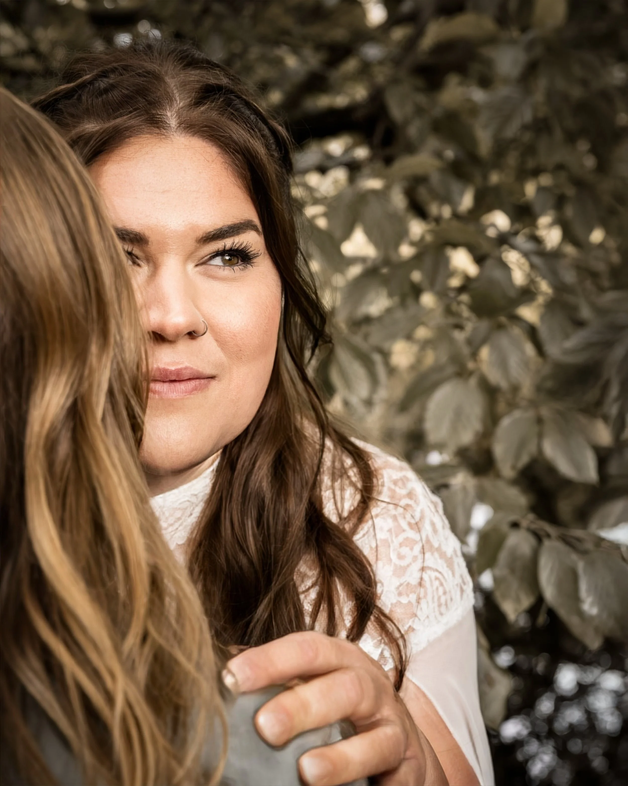 Zwei Frauen umarmen sich, eine davon ist sichtbar im Bild, mit braunen Haaren, im Hintergrund eine Büschel von Blättern oder Bäumen.