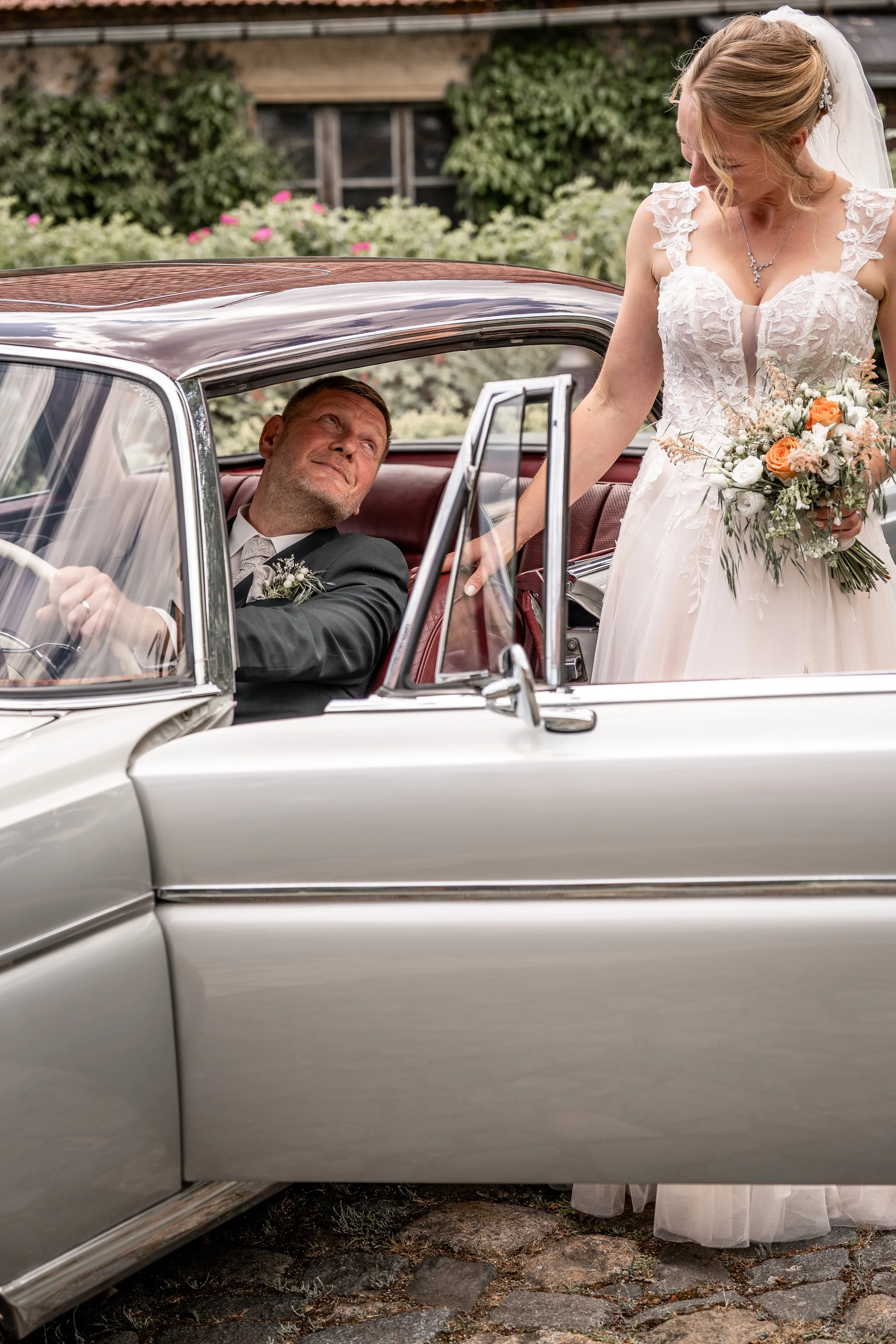 Braut in Hochzeitskleid steht neben einem klassischen weißen Auto im Freien.