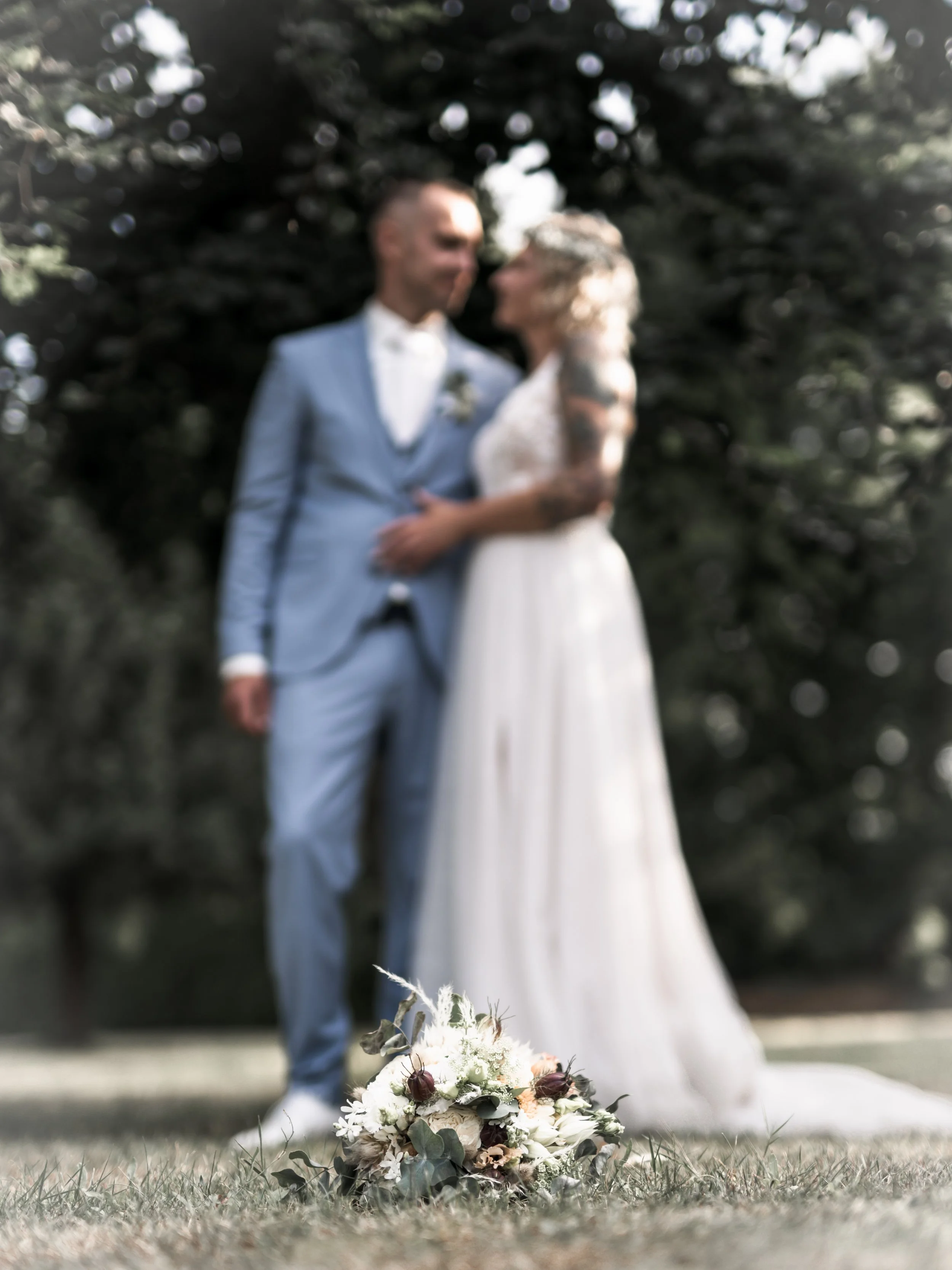 Ein Brautpaar bei einer Hochzeit im Park, mit einem Brautstrauß auf dem Rasen vor ihnen, im Hintergrund Bäume und Himmel.