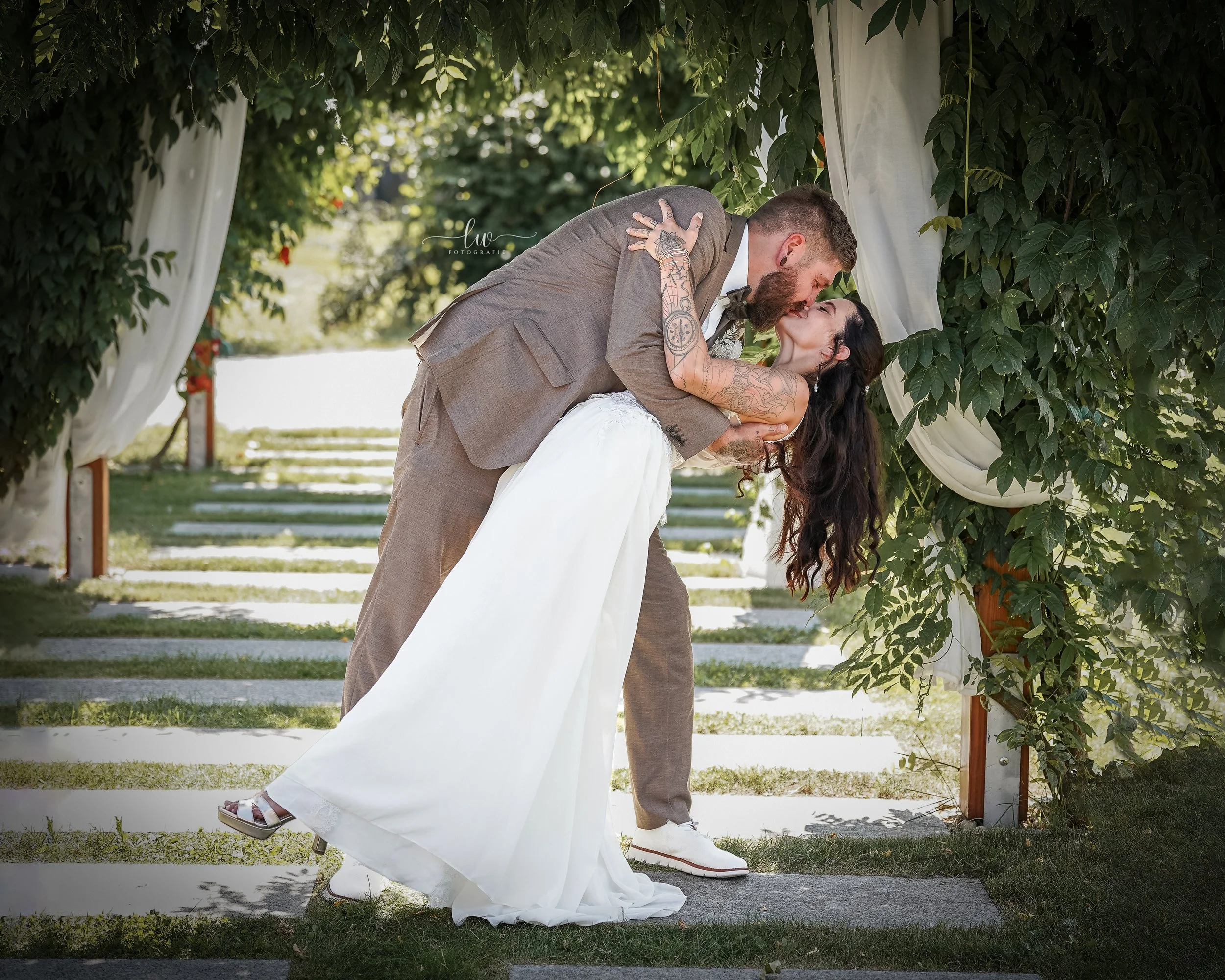 Ein Paar bei ihrer Hochzeit, das sich küssen, während sie unter einem Bogen aus grünen Blättern und weißen Tüchern im Freien steht.
