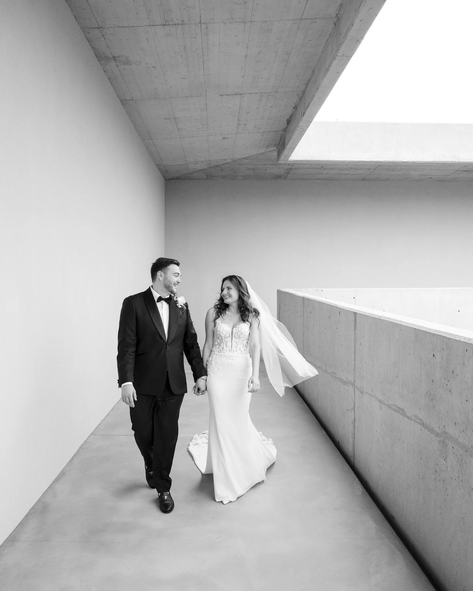 Black and white photo of a bride and groom walking hand in hand in a modern architectural setting, smiling at each other.